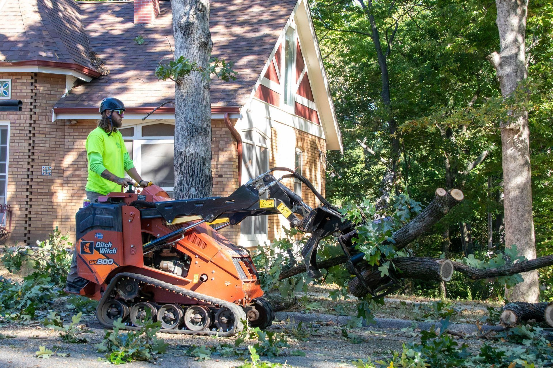 A person uses a mini skid steer with a saw to cut a tree branch near a house.