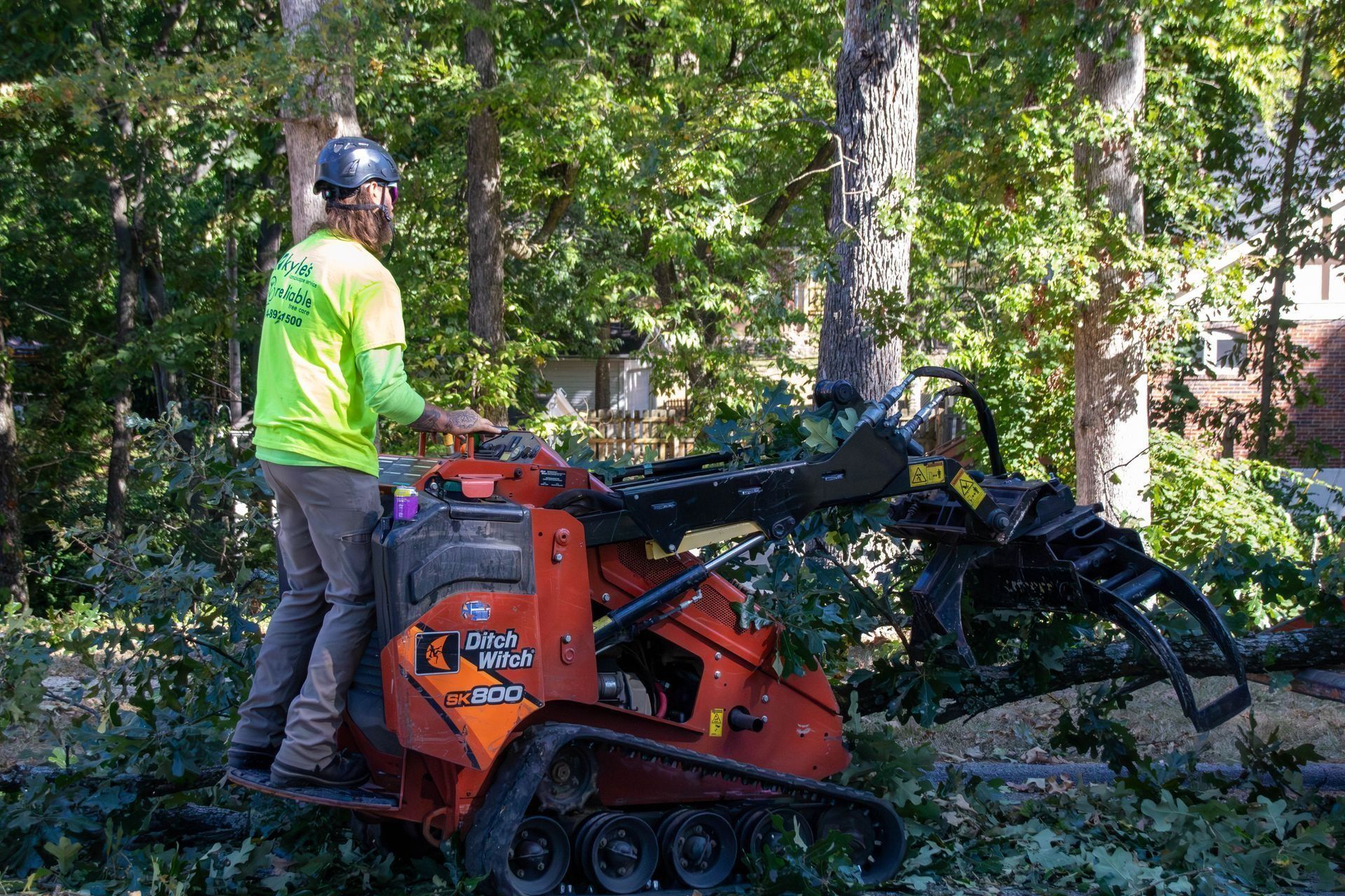 Person operating an orange mini-excavator with a claw attachment, cutting branches. Outdoors in a wooded area.