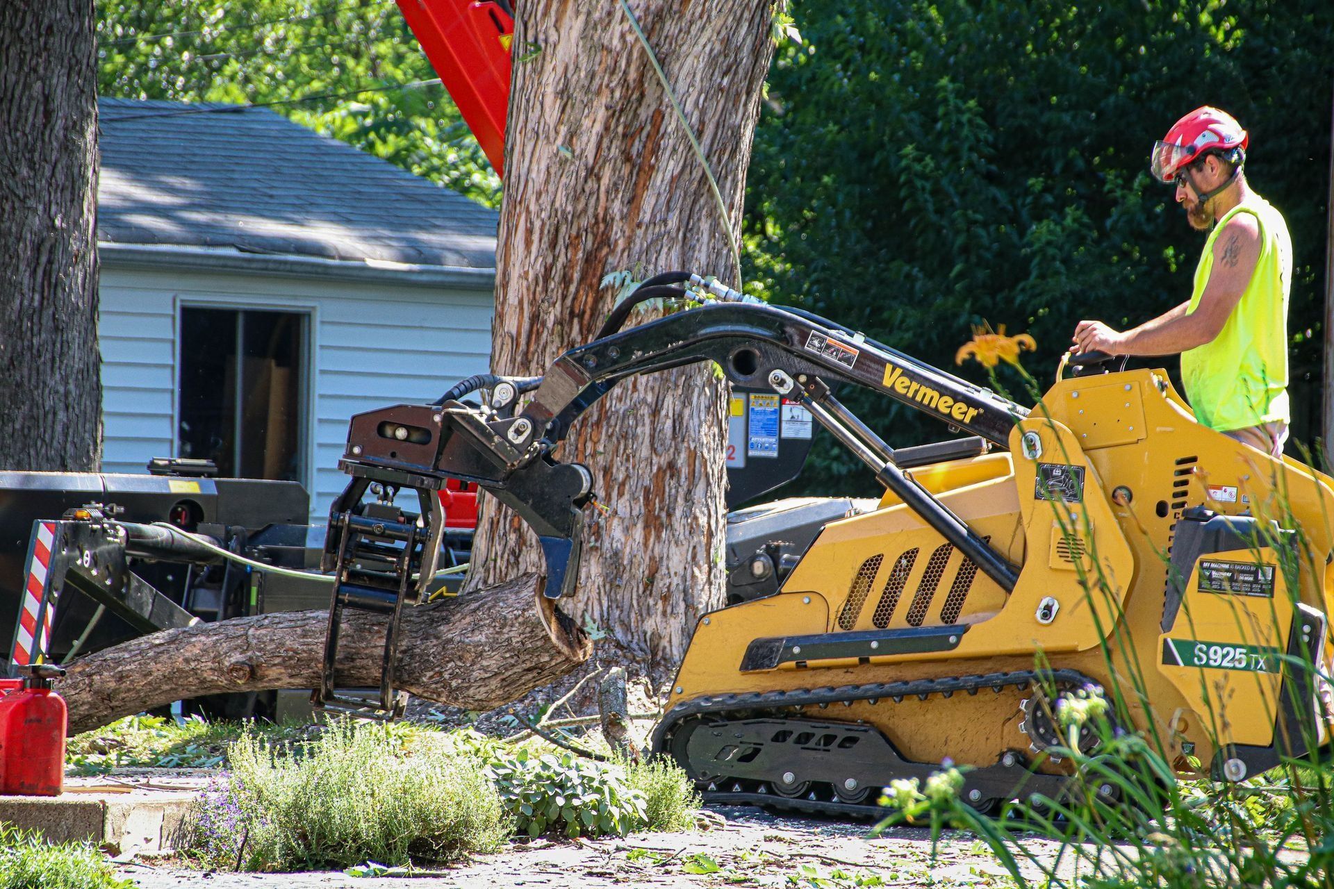 Man operating a yellow skid steer, cutting a tree trunk. Residential setting with small house visible.