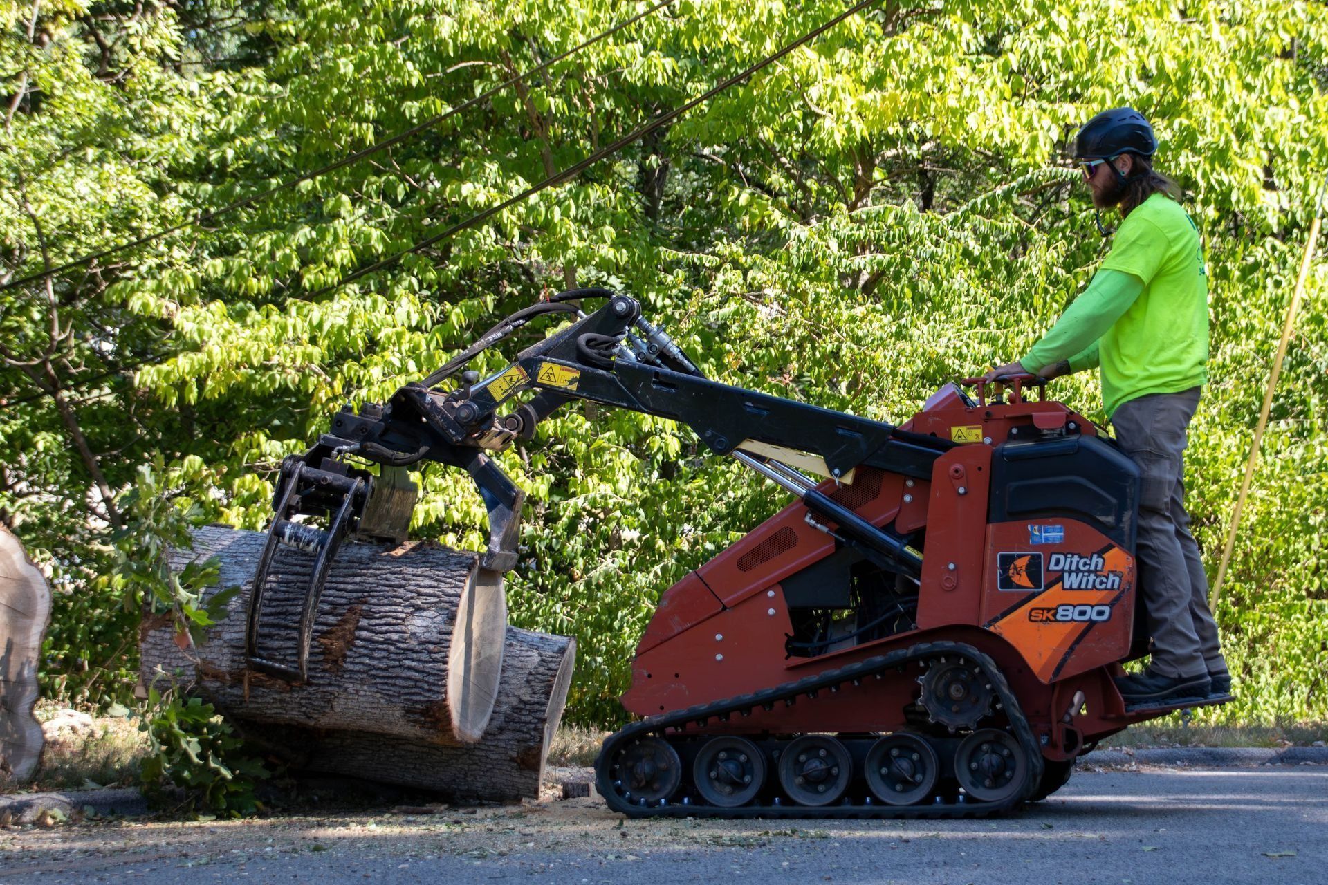 Man operating a small, orange tracked machine with a log-grabbing attachment, lifting a log in a wooded area.