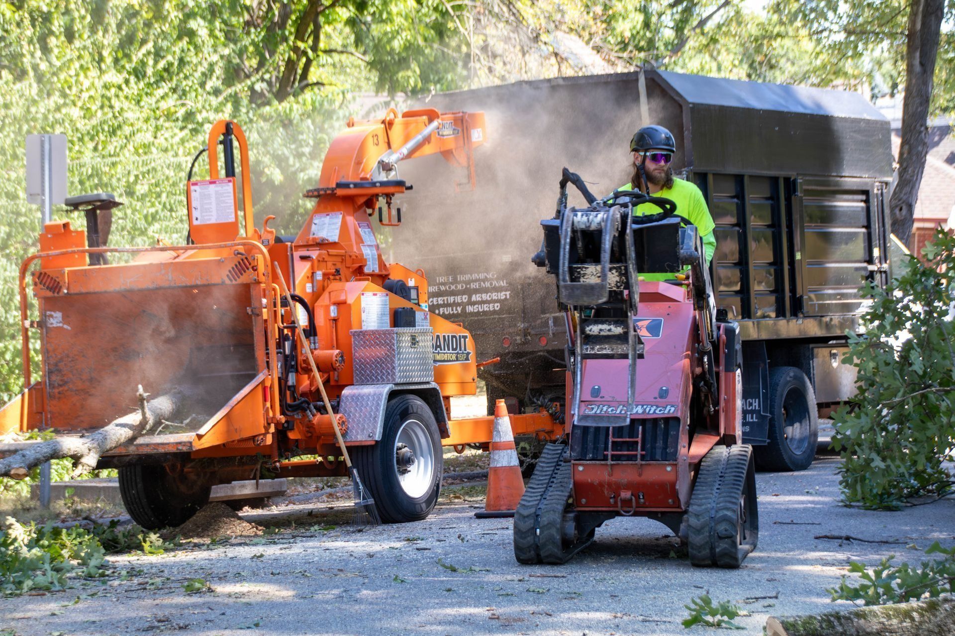 Man operating wood chipper, chips blowing into truck; tree removal activity.