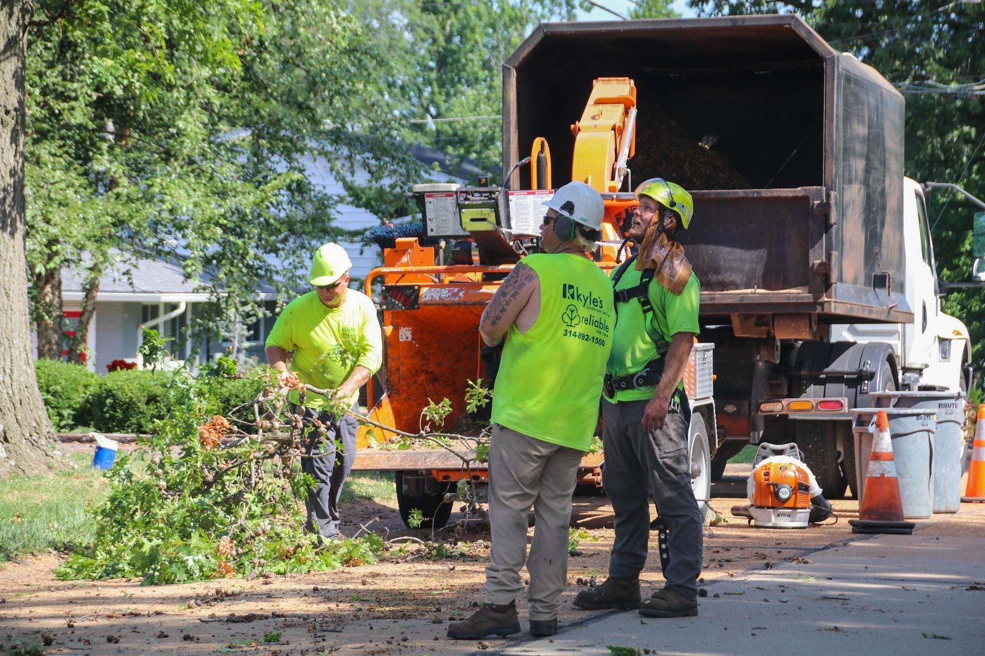 Three workers in neon green vests, helmets, and work clothes; chipping tree branches next to truck on a street.
