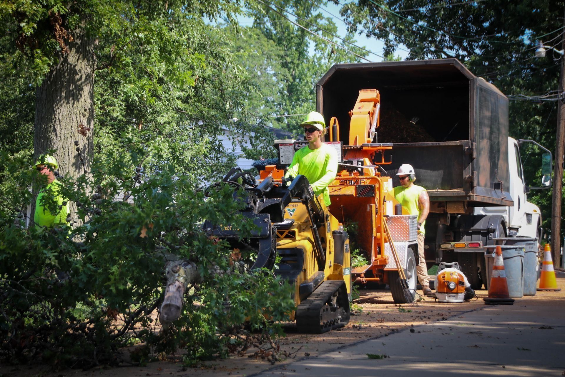 Tree trimmers in safety vests using a wood chipper on a street with a truck parked nearby.