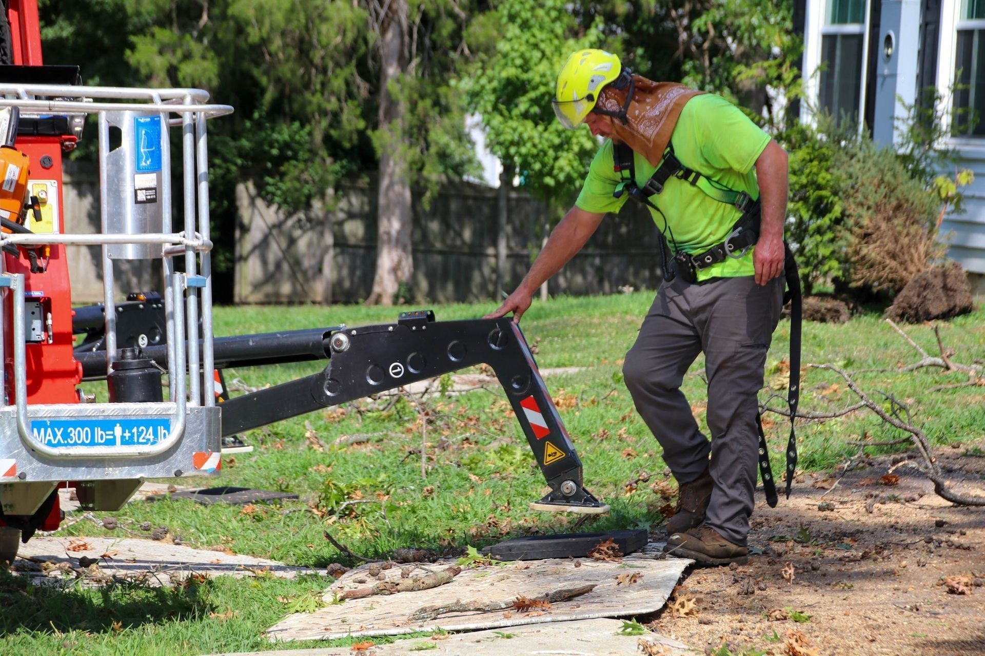 Man in safety gear near a cherry picker, adjusting a support arm on a grassy lawn.