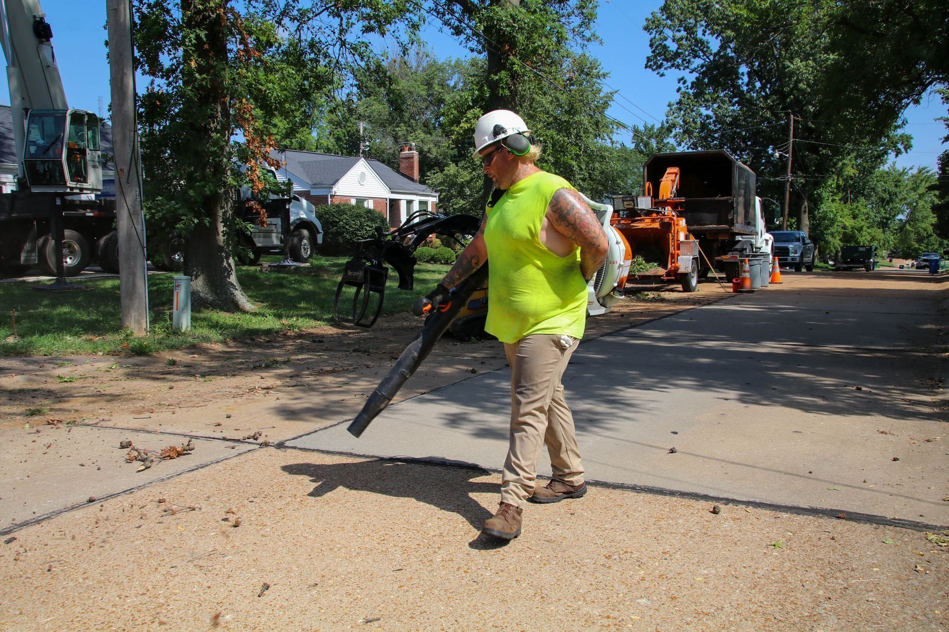 Man in safety vest blowing debris on a street, with tree trimming equipment in the background.