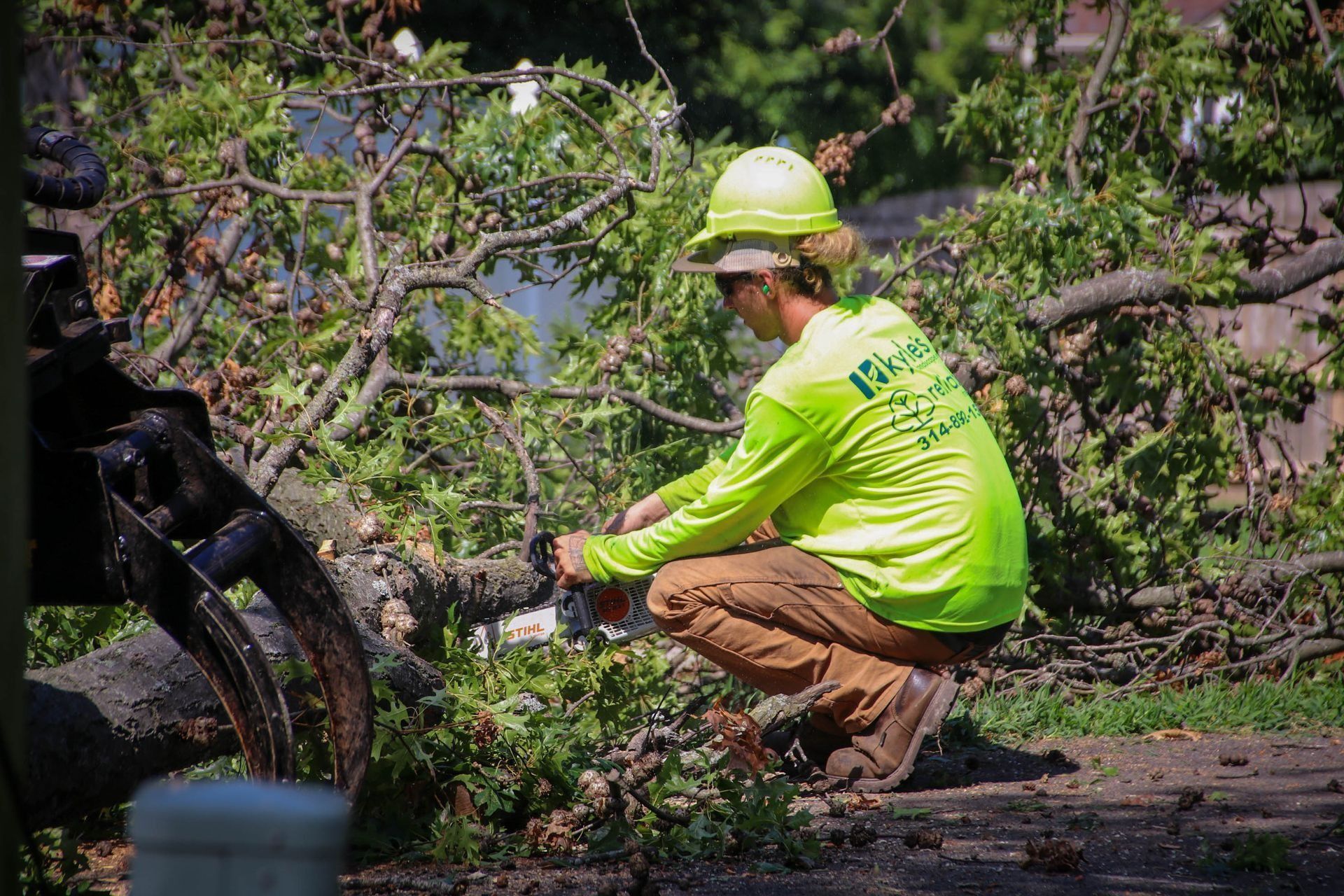 Tree service worker using chainsaw on a fallen tree branch. Person wearing neon green long sleeve, hard hat, and brown pants.