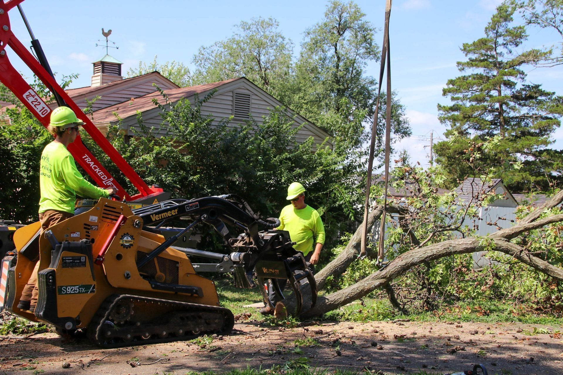 Two workers use heavy machinery to remove a fallen tree in front of a house.
