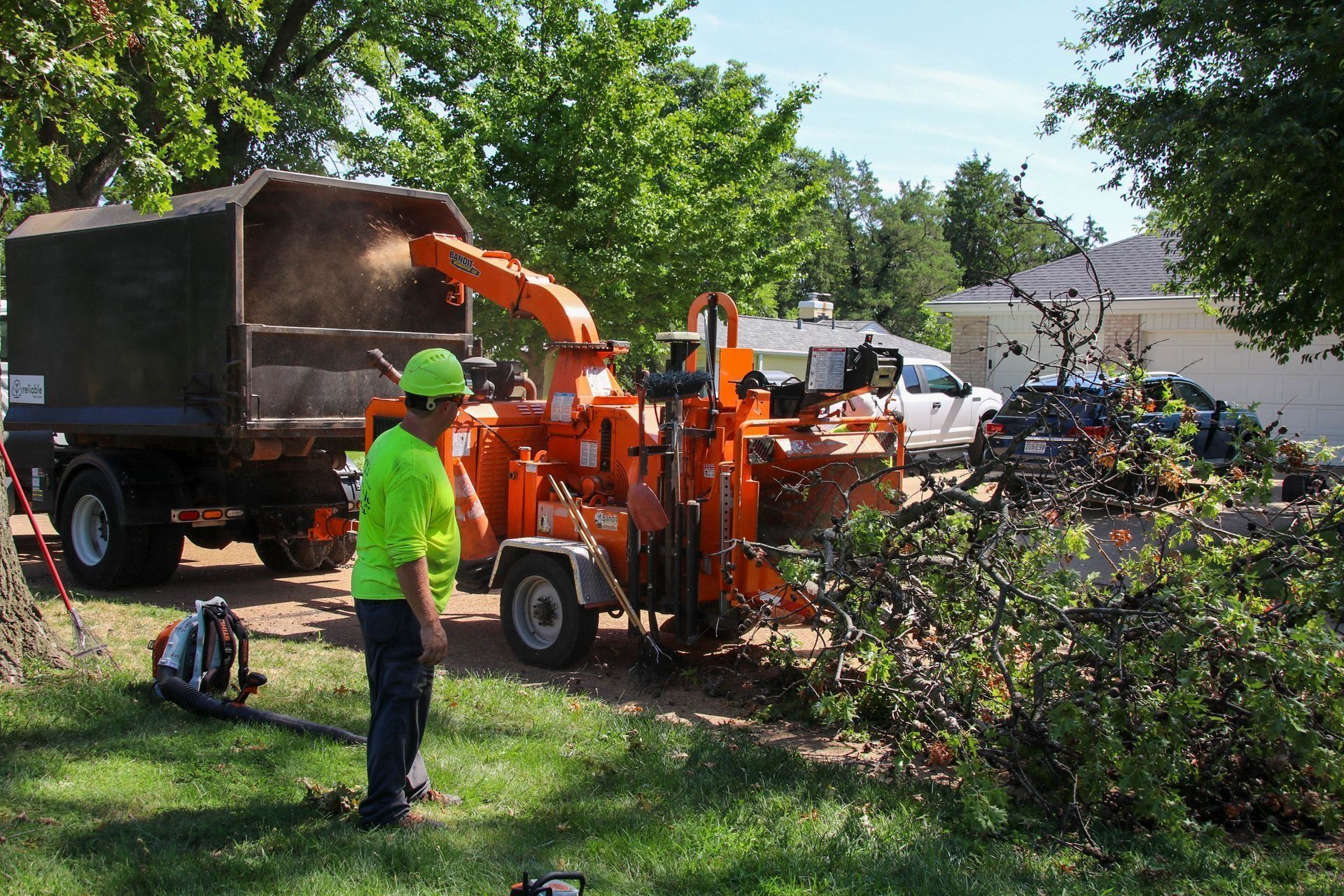 Tree chipper processing branches, worker in safety gear, truck, residential yard setting.