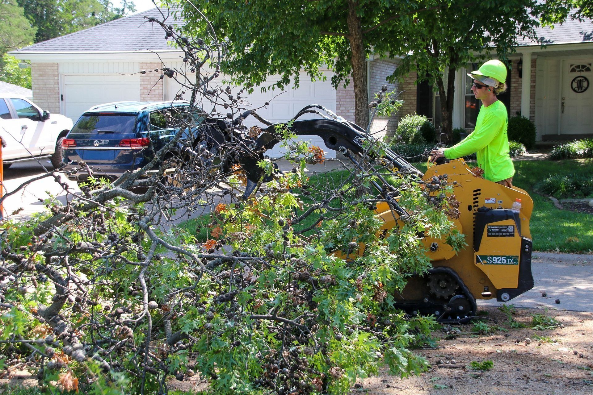 Man operating a skid steer, removing tree branches on a residential street.