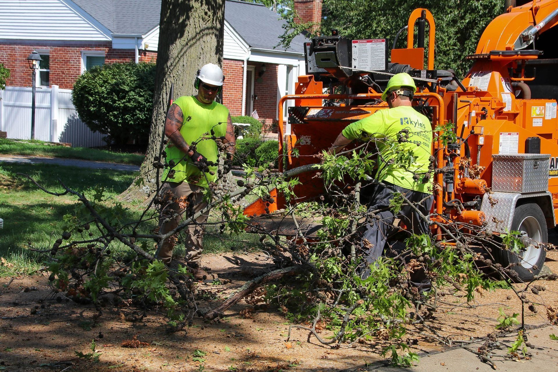 Two workers in safety vests feed tree branches into a wood chipper in a residential area.