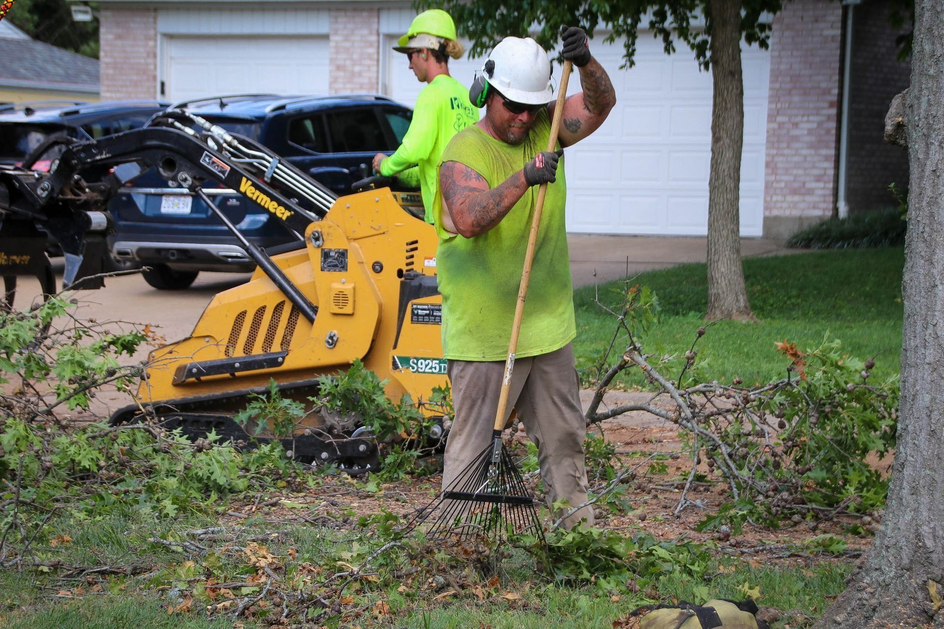 Two workers raking debris near a yellow skid steer. They wear safety gear outside a house with a car.