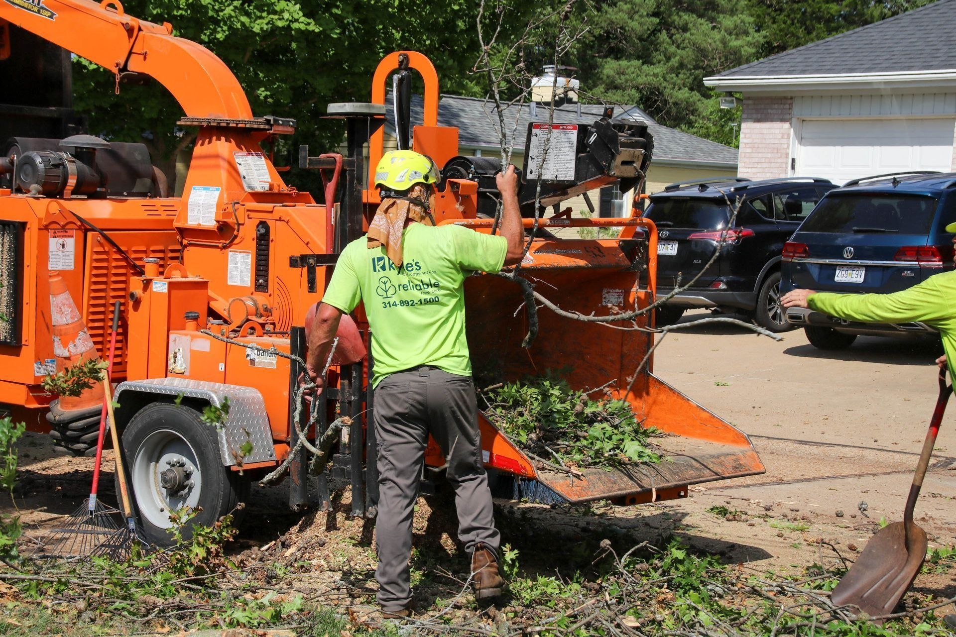 Worker feeding tree branches into an orange wood chipper; house and cars in the background.