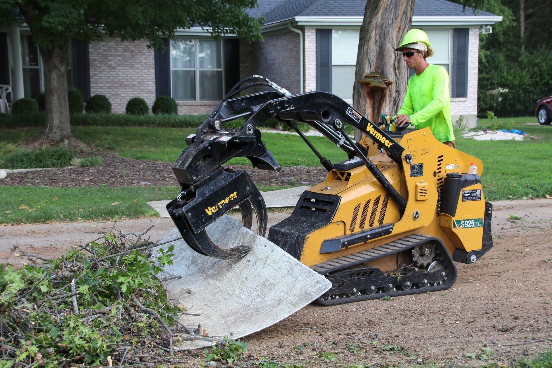Person operating a yellow mini-skid steer with a grapple attachment, moving debris on a gravel road in front of a house.