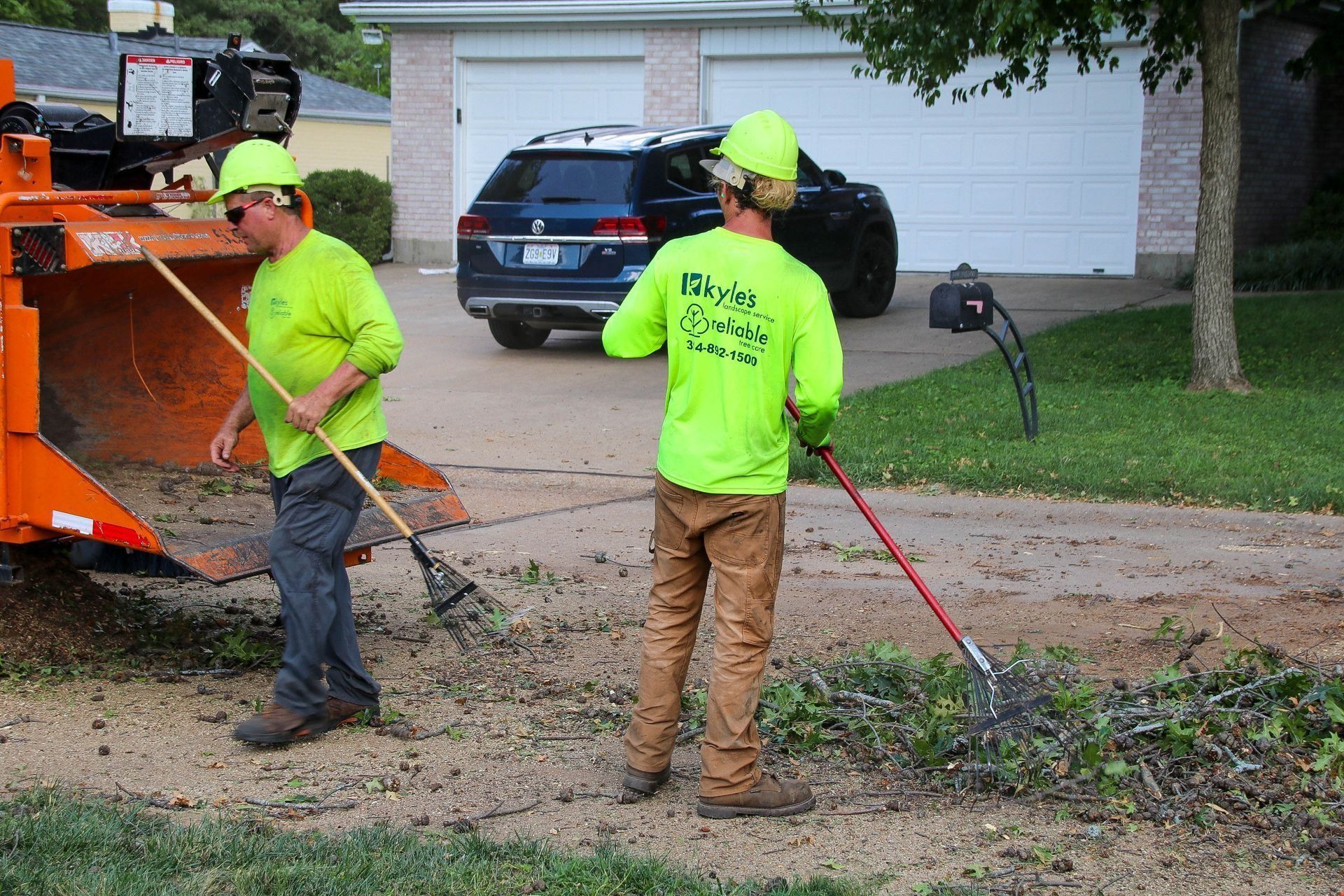 Two workers in neon green shirts rake debris near a wood chipper in a driveway.