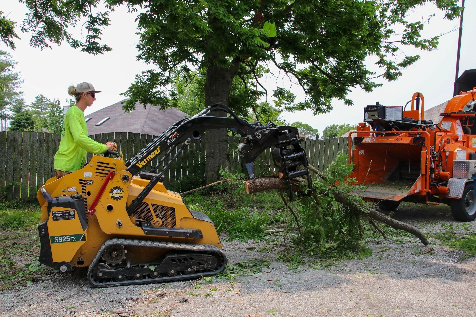 A person operating a yellow compact excavator with a grapple arm, feeding tree branches into a wood chipper.
