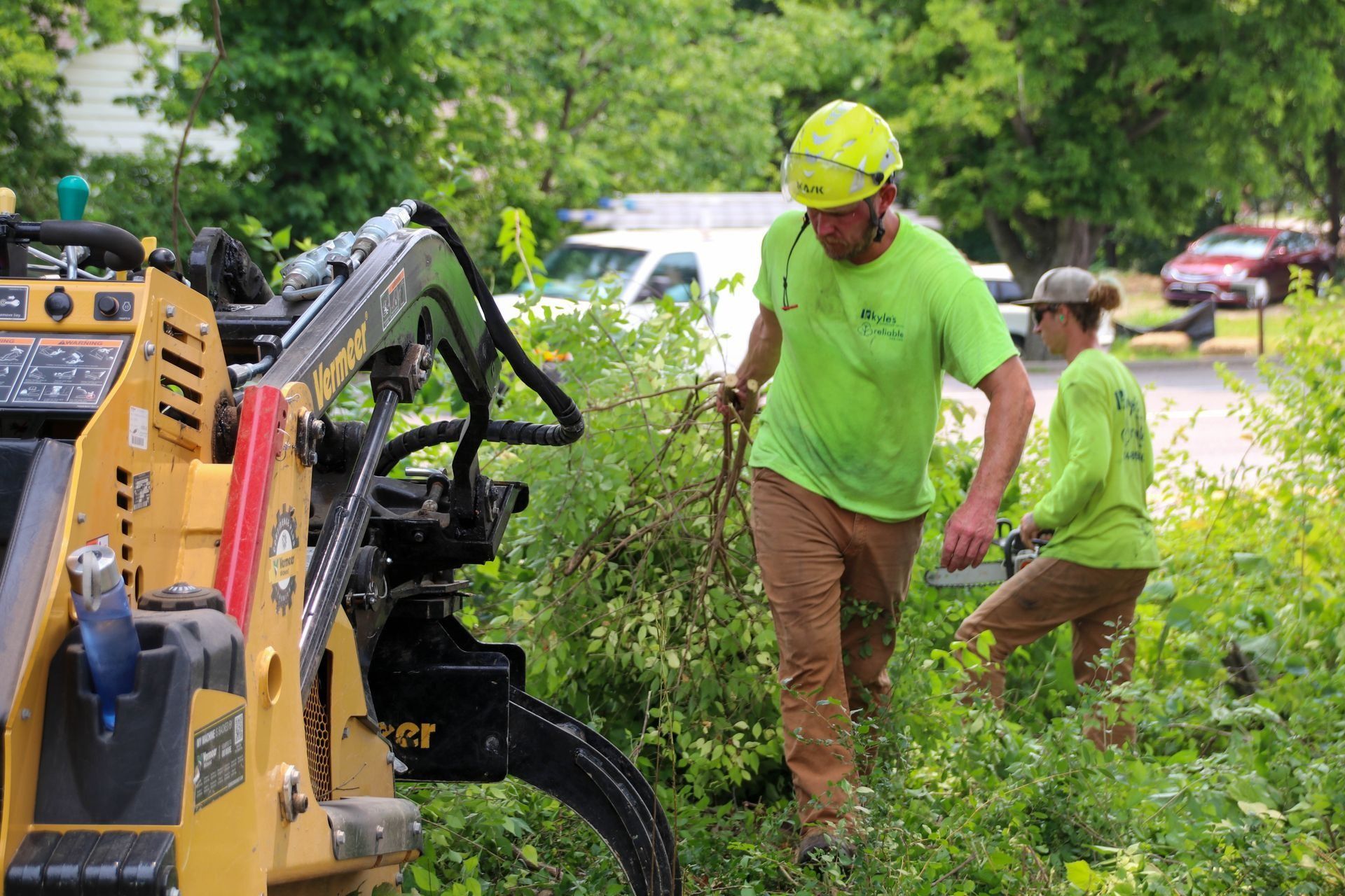 Two workers in green shirts and hard hats trimming bushes with a machine and tools.