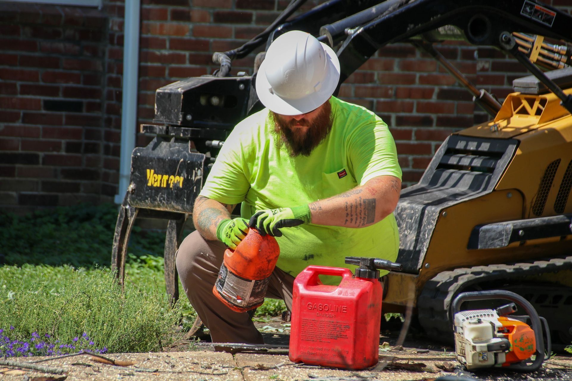 Construction worker in white hard hat and green shirt pouring fuel from a red gas can.