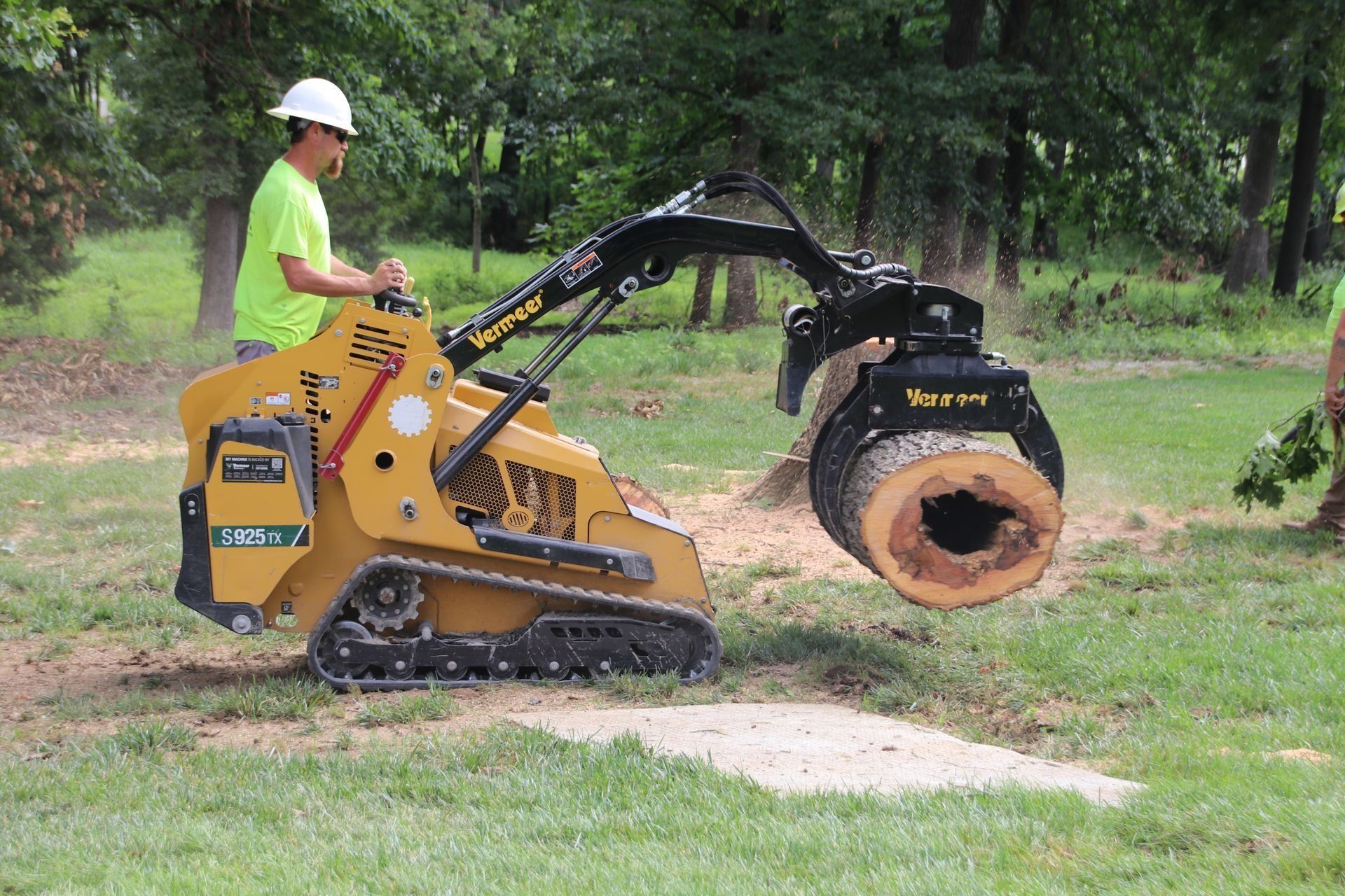 Man operating a yellow skid steer with a log grapple, lifting a tree trunk. Green field setting.