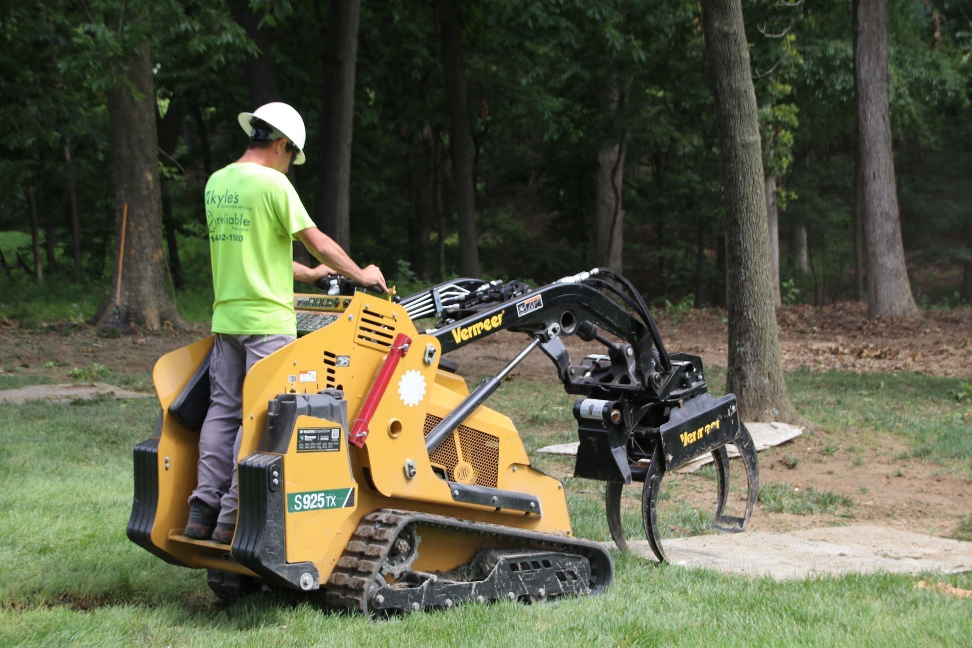 Man operating a yellow skid steer with tree stump grinder in a grassy area near trees.