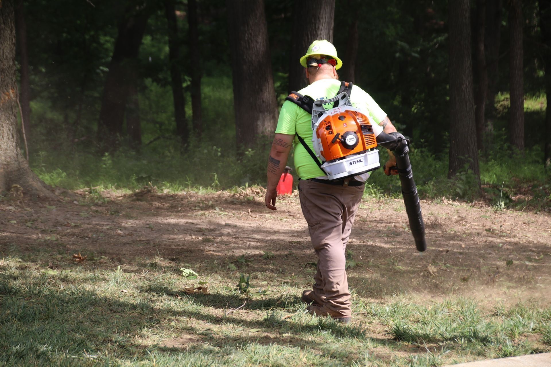 Person with backpack blower walking on grass; trees in background.