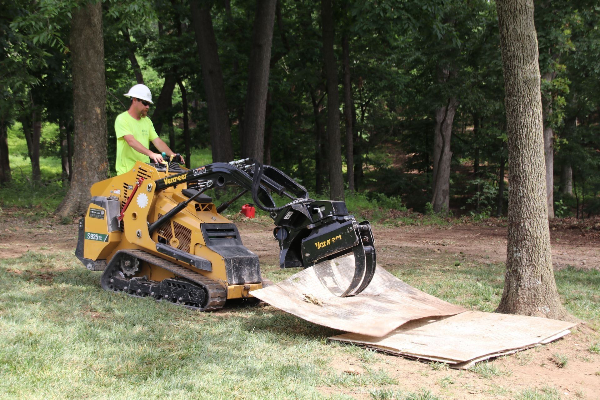 Man operating a compact track loader with grapple attachment in a wooded area, lifting cardboard.