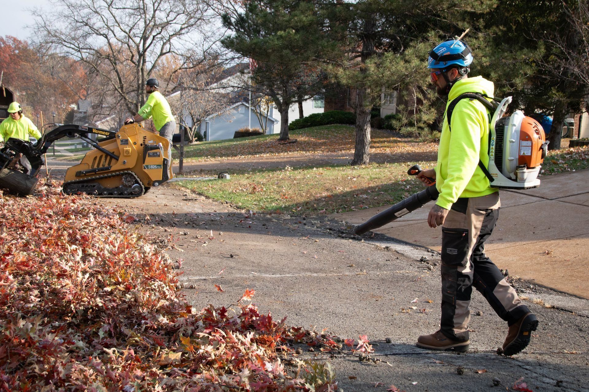 Three workers in neon green clothing clearing leaves from a street with a leaf blower and a skid steer.