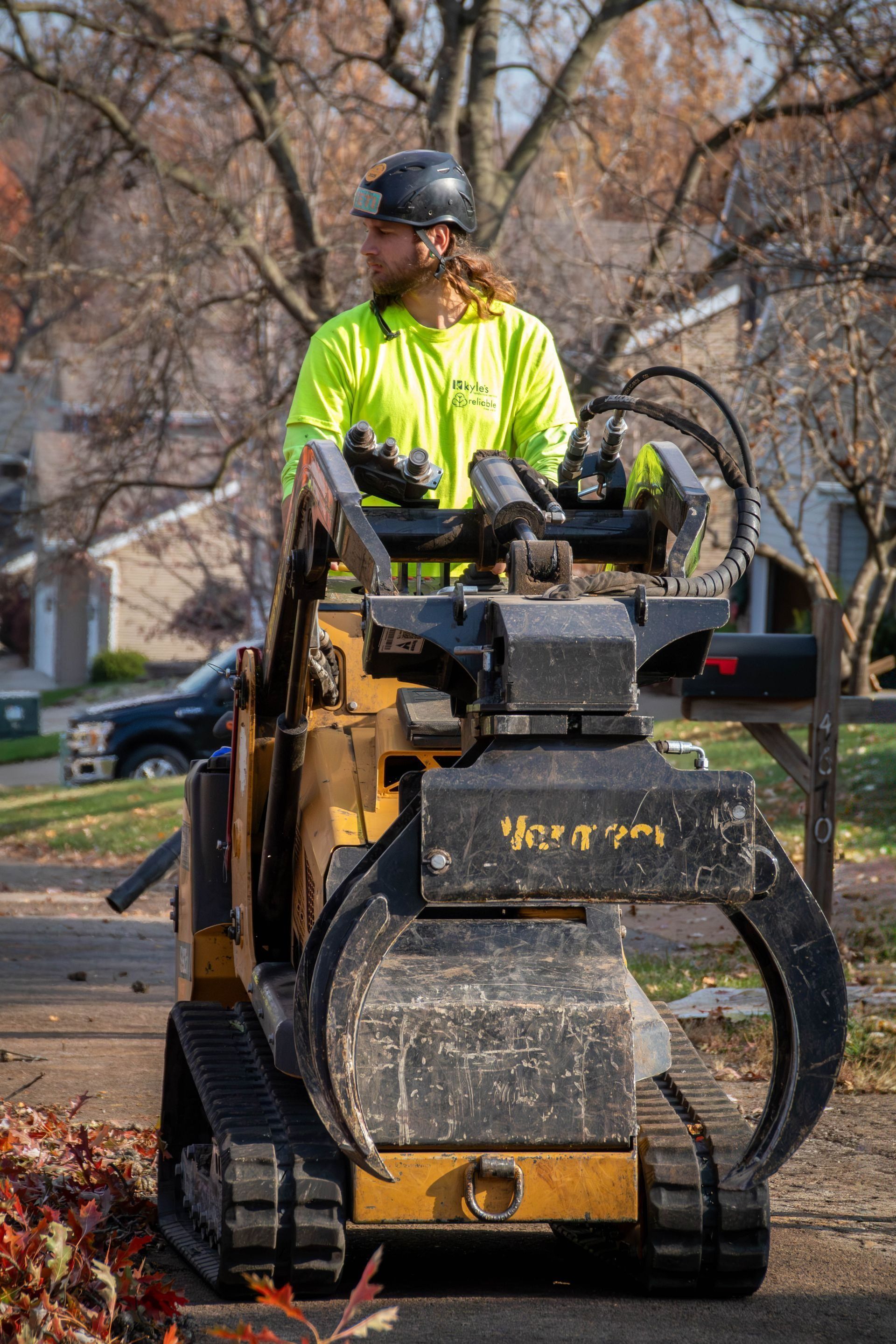 Man operating a small skid-steer loader with grapple attachment in a residential area. Yellow safety shirt.