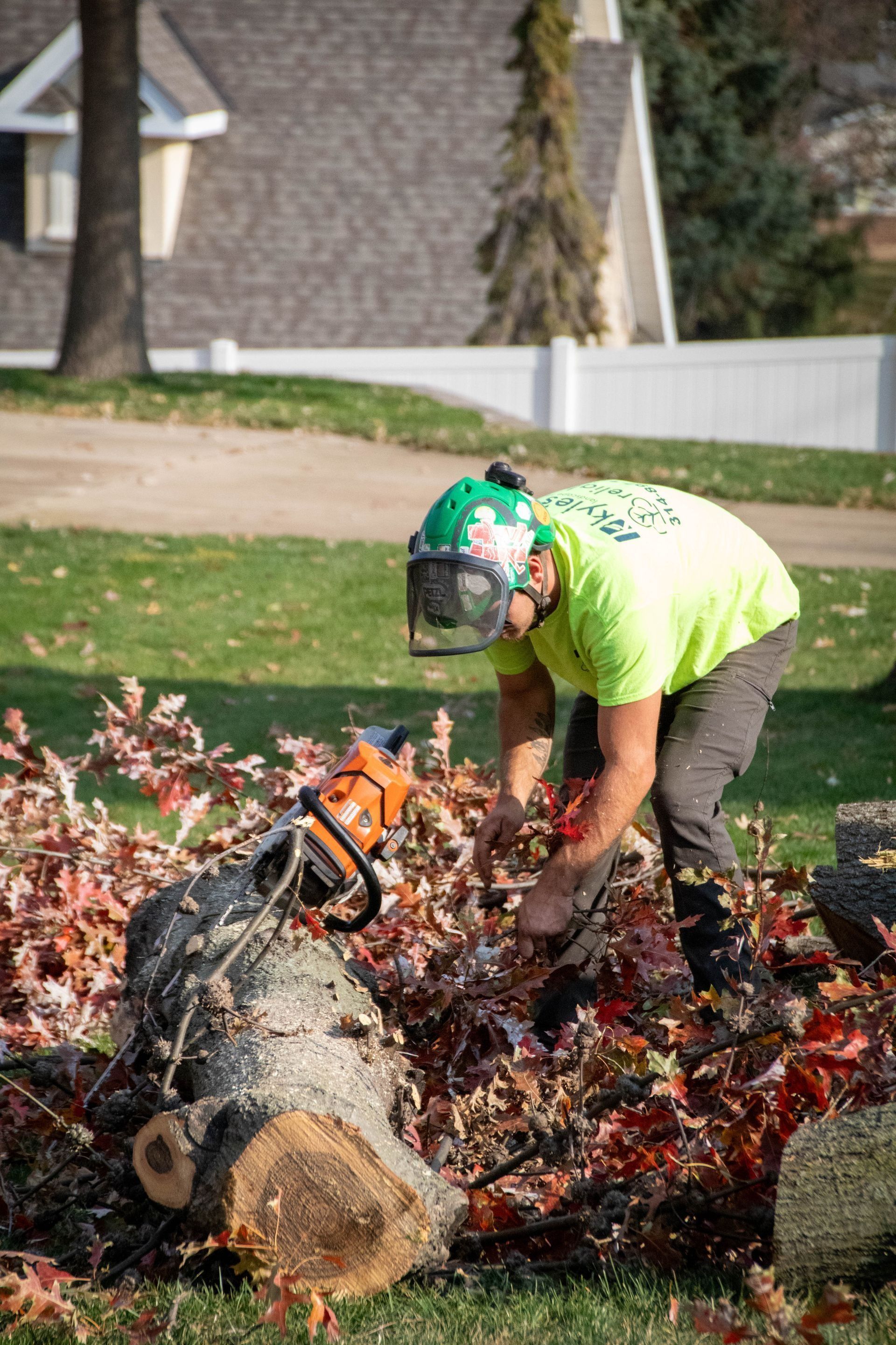 A person wearing safety gear uses a chainsaw to cut a log on a lawn, surrounded by debris.