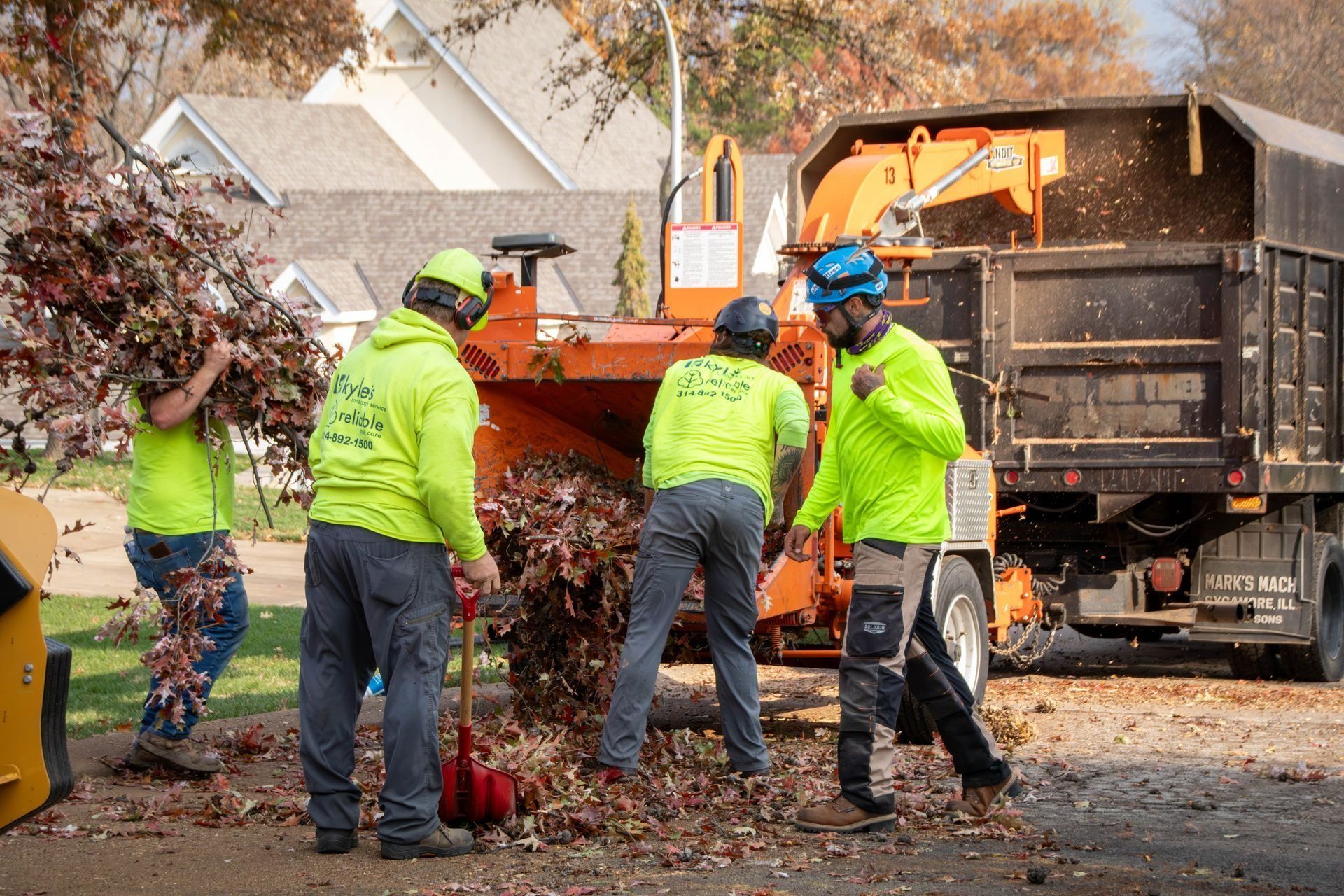 Tree service workers using a wood chipper to shred branches; truck nearby.