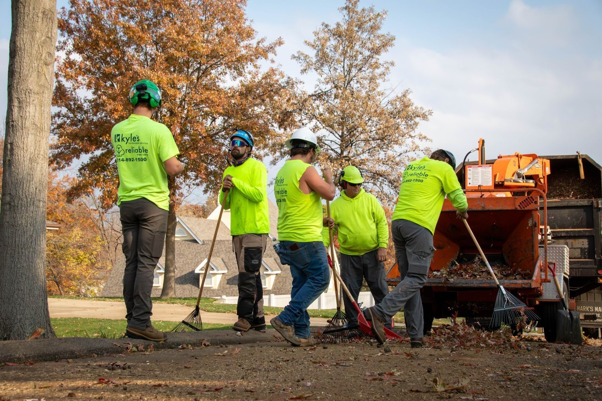 Five workers in neon green shirts rake leaves near a wood chipper and a tree in a residential area.