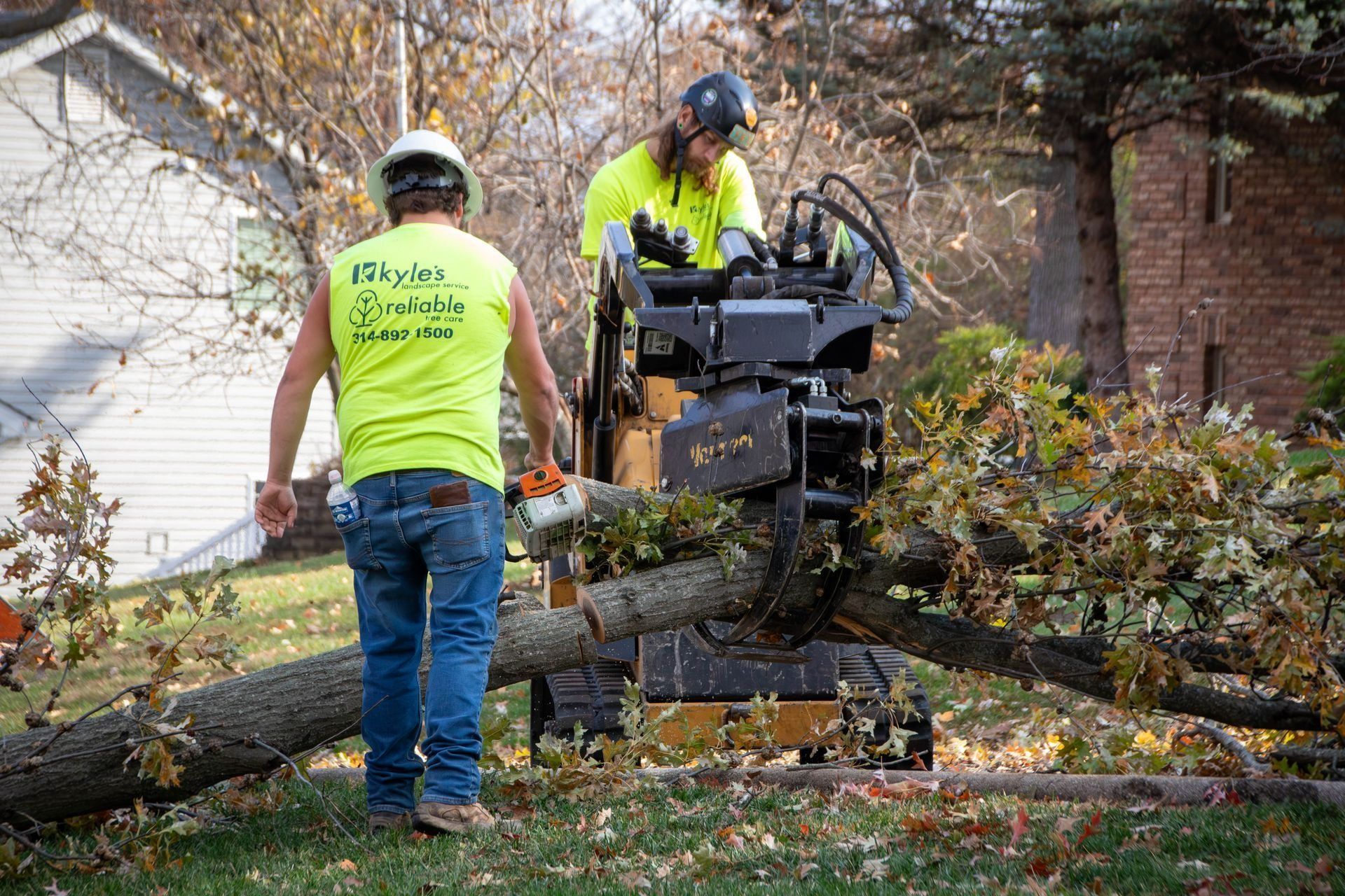 Two workers use machinery to cut a fallen tree limb on a grassy lawn.