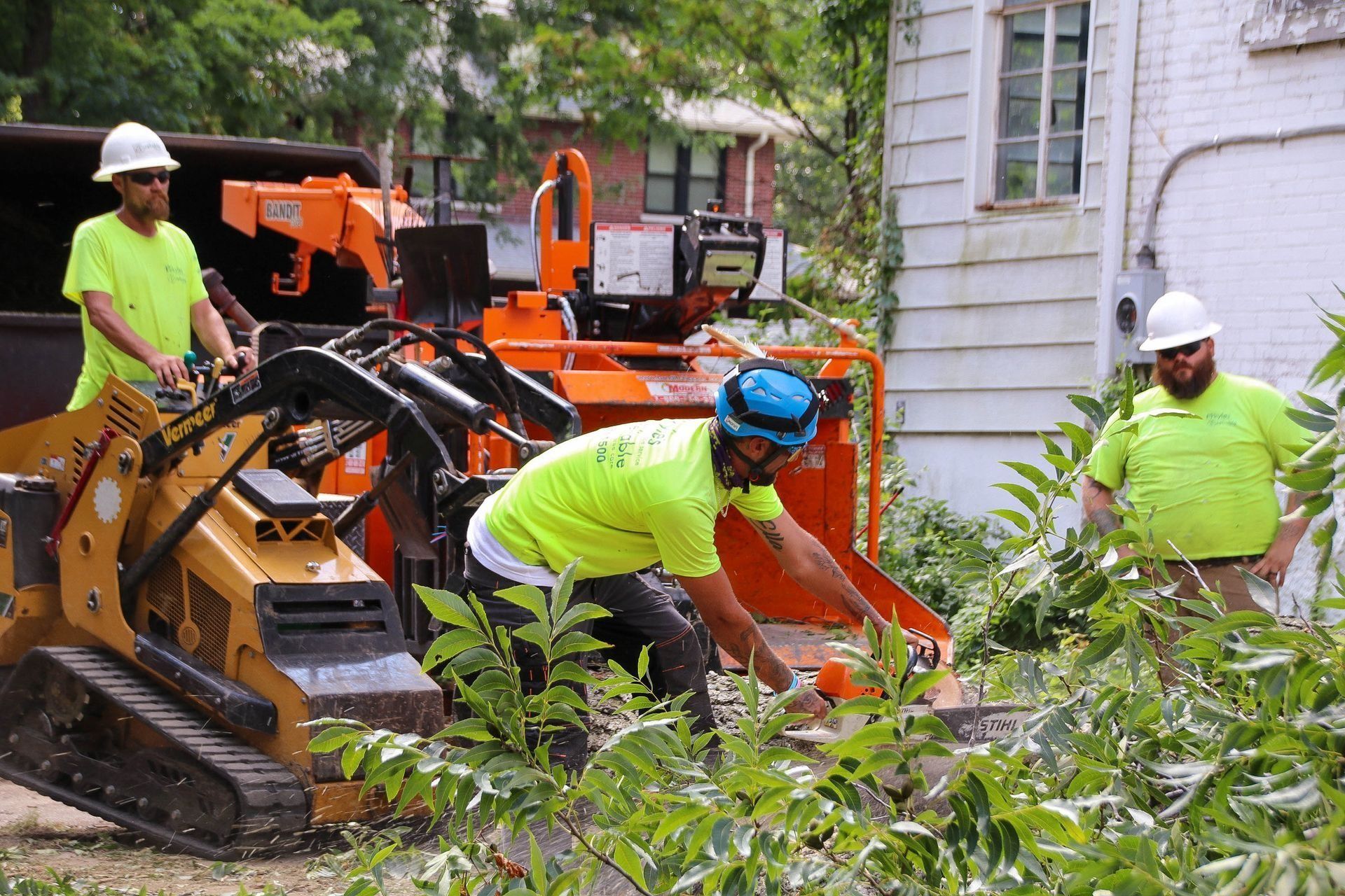 Three people in safety gear operating a wood chipper near a house.