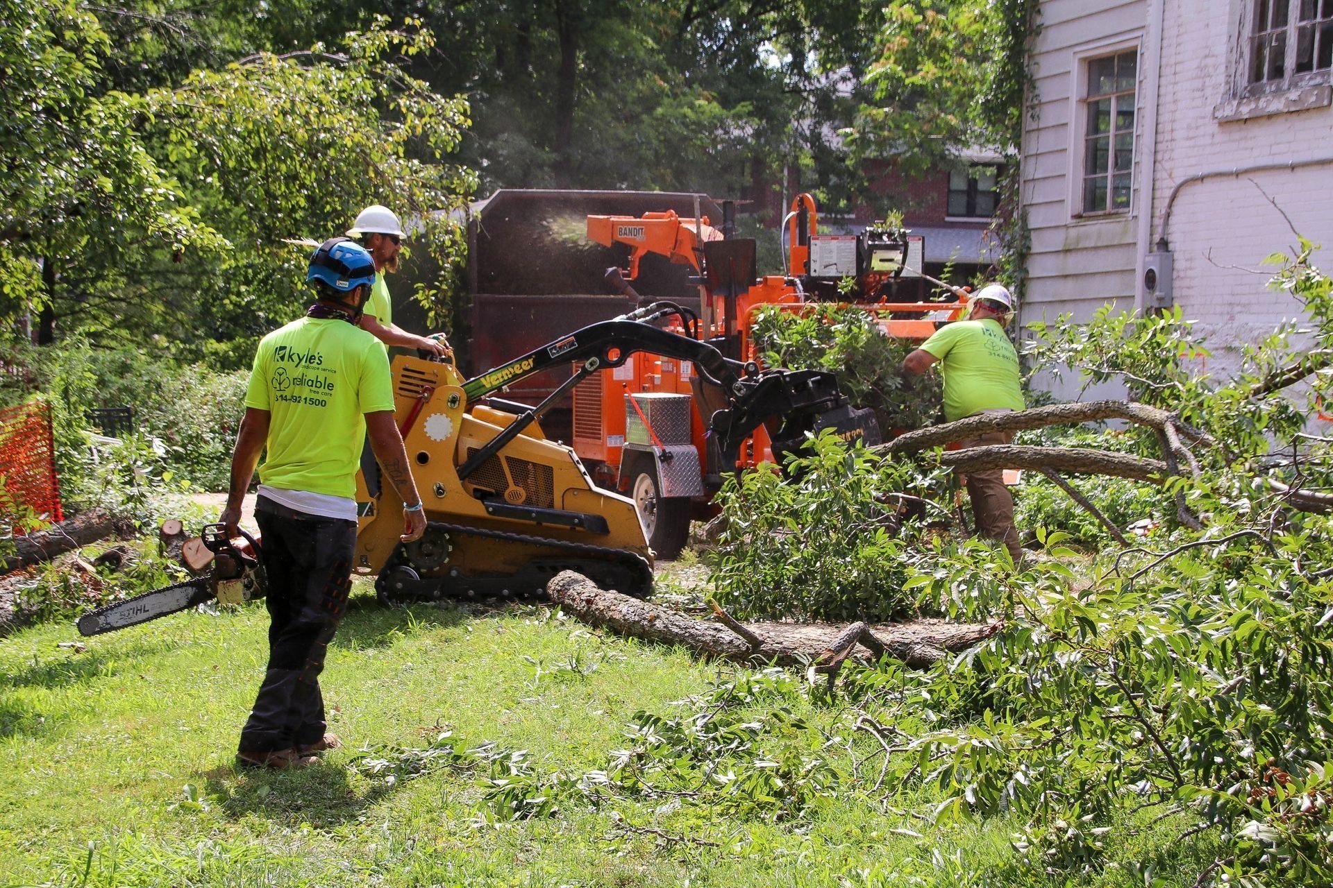 Tree service crew using a chipper. Men in safety vests work with branches near a house and machine.