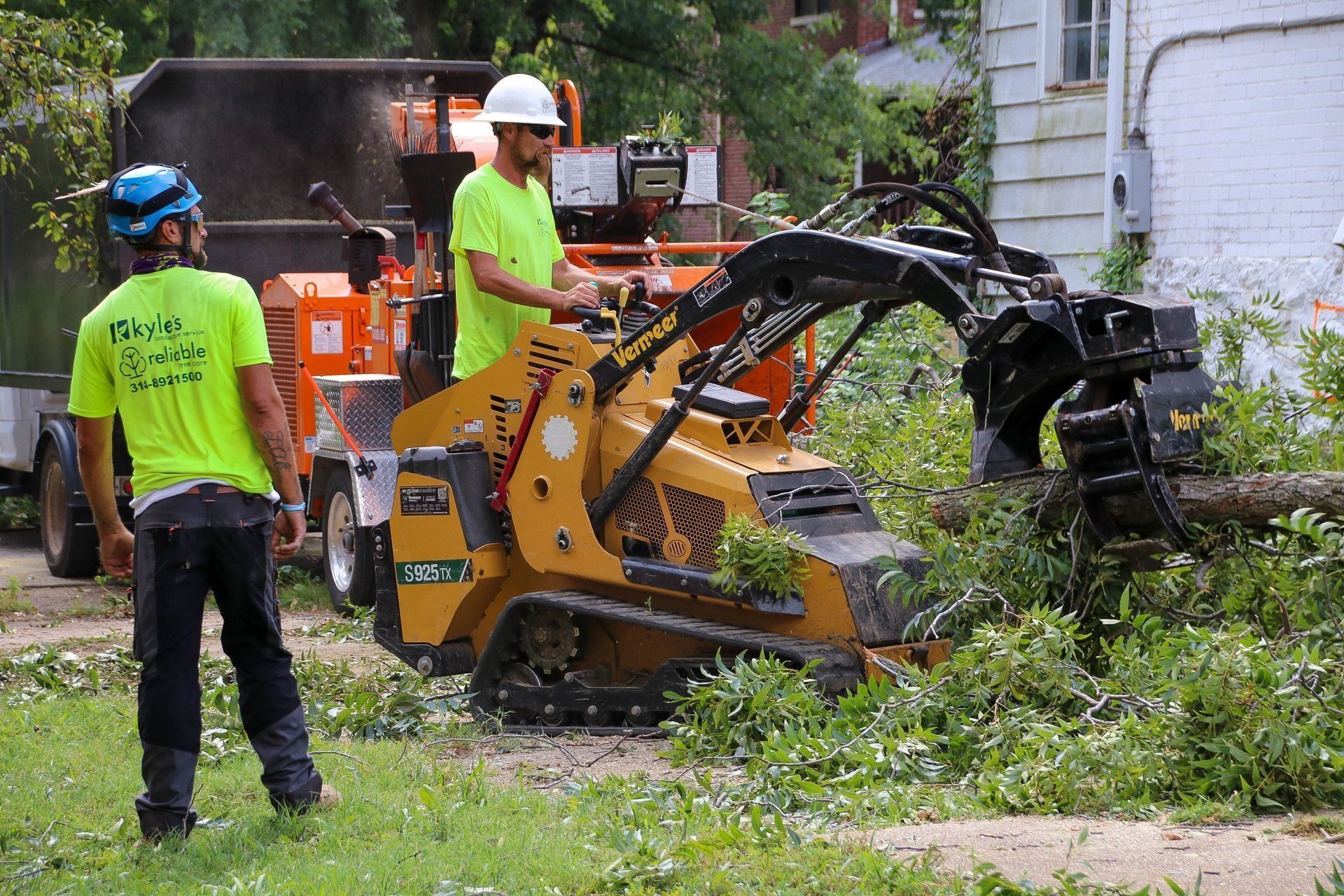 Two workers using a skid steer with a grapple to move branches. One worker operates a wood chipper. Outdoors.