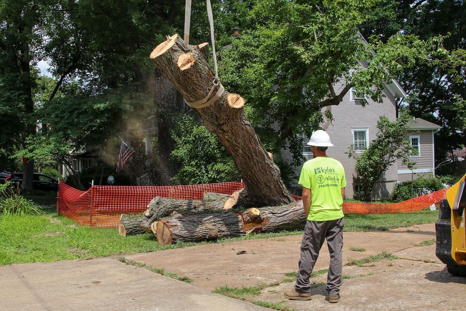 Tree removal: worker in safety gear watches crane lifting a cut tree section. Orange barrier, suburban setting.