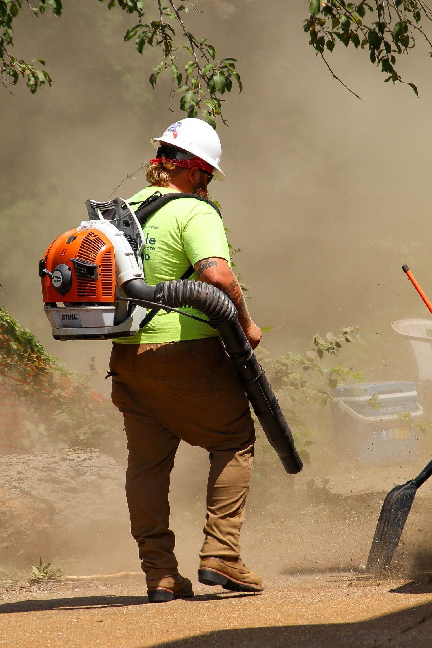 Person using a leaf blower, kicking up dust. They wear a helmet, green shirt, and brown pants, standing outdoors.