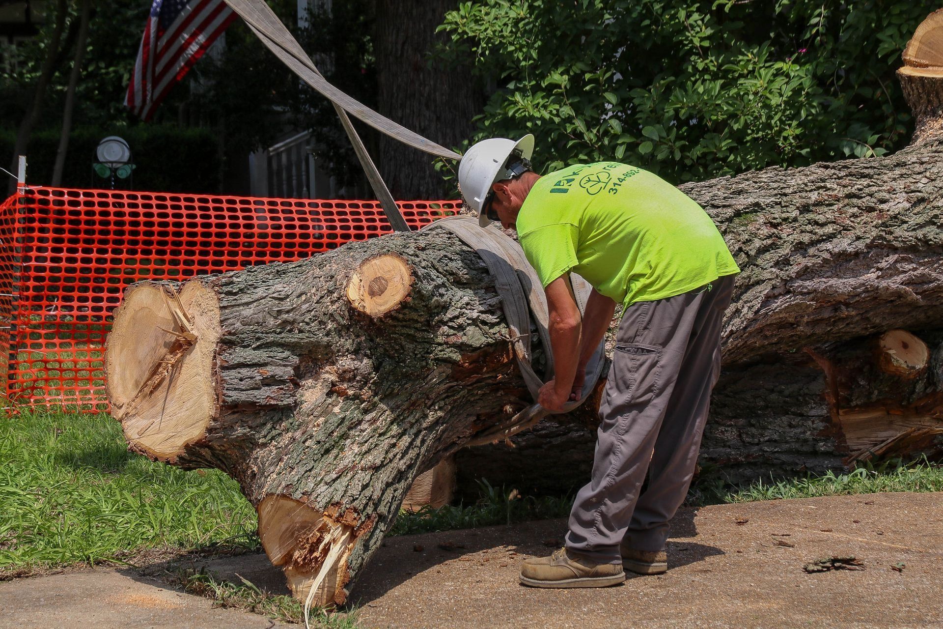 Man in neon shirt sawing a large fallen tree trunk. Orange fence and American flag in background.