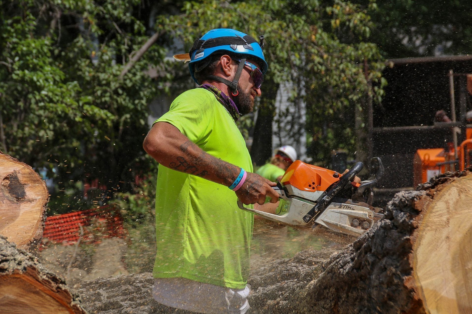 Man in neon shirt using a chainsaw to cut a log, wearing a blue helmet and safety glasses.