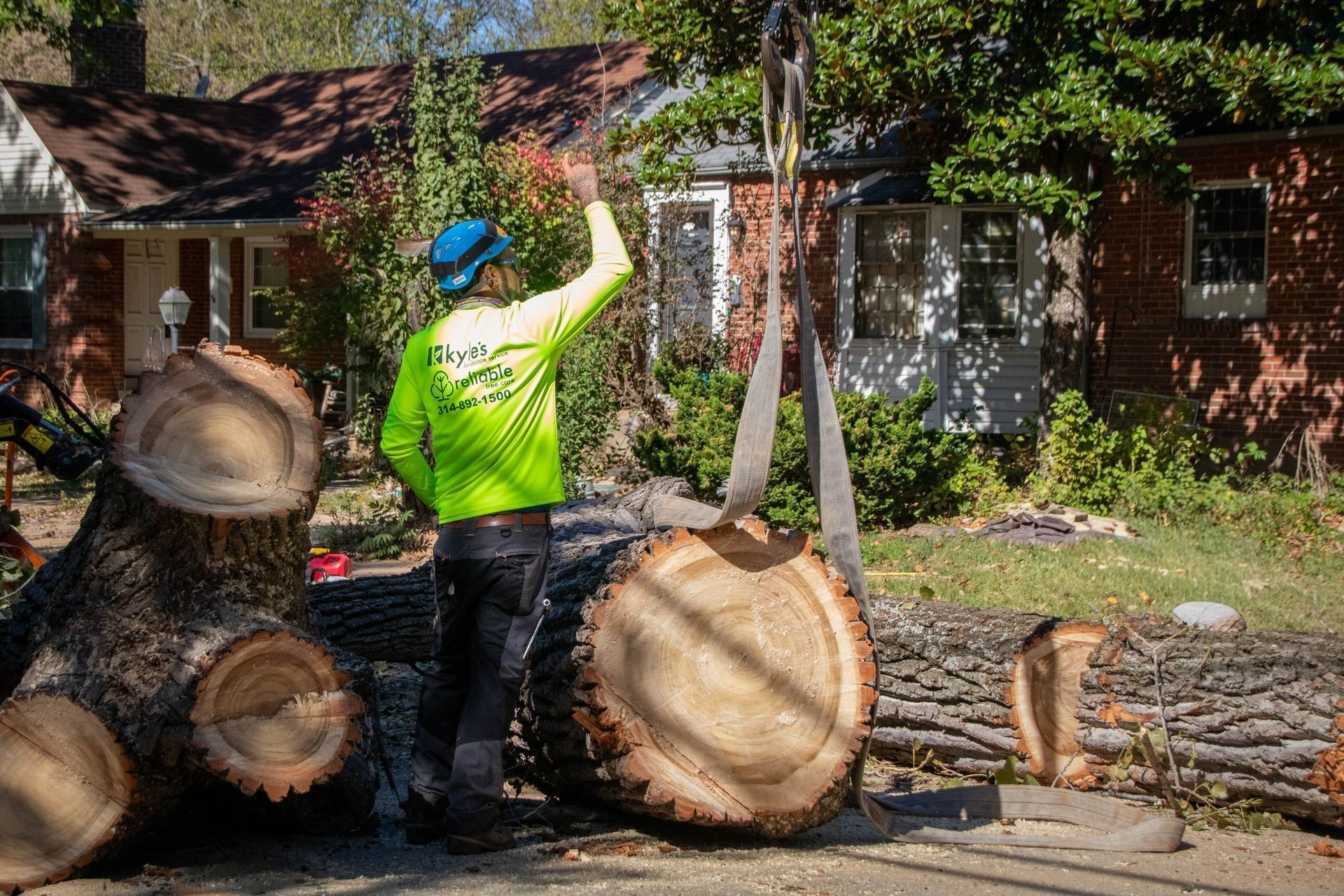 Tree service worker in neon shirt directs lifting of a large tree trunk. House in background.