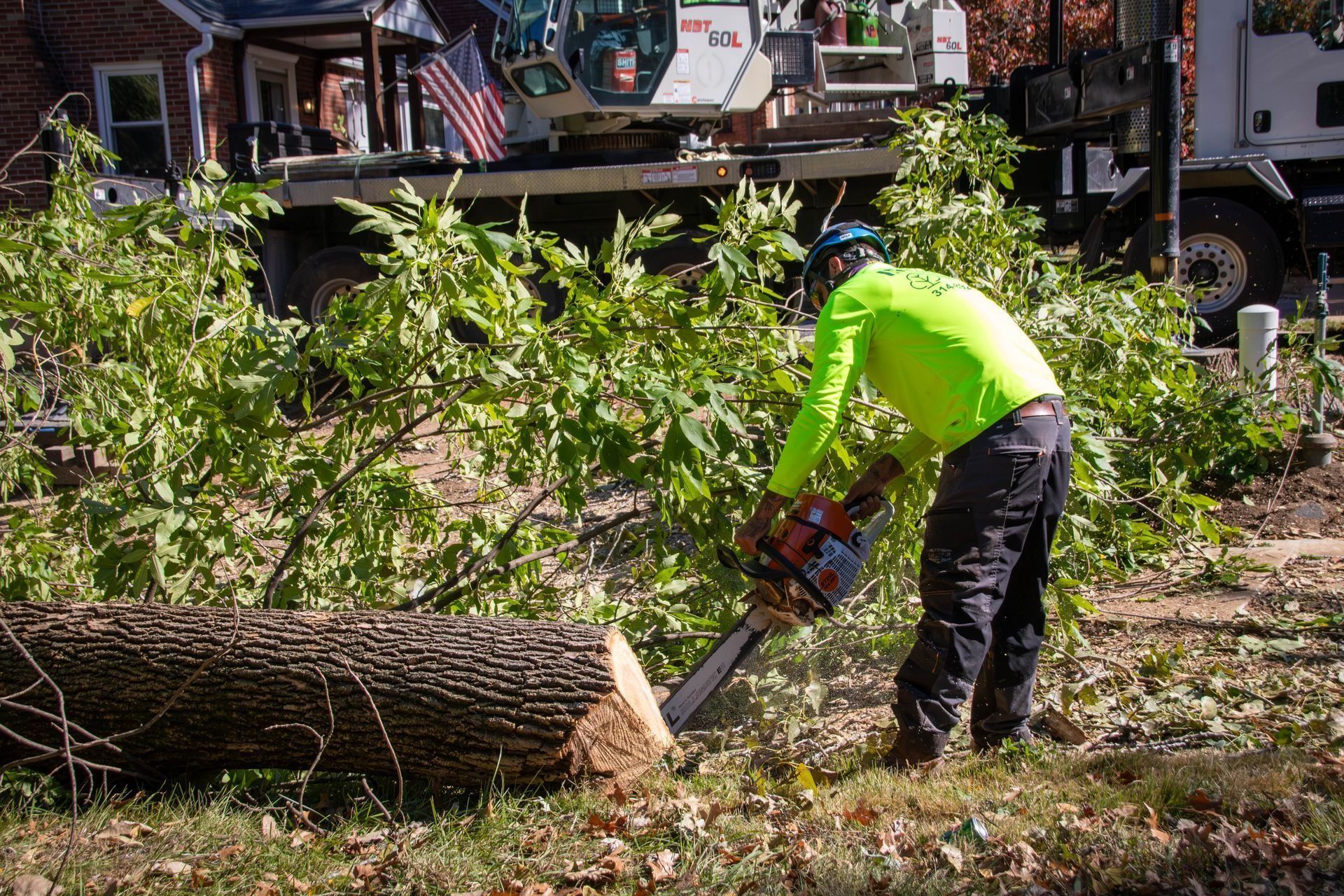 Arborist cutting log with chainsaw; green shirt, safety gear, crane truck in background.