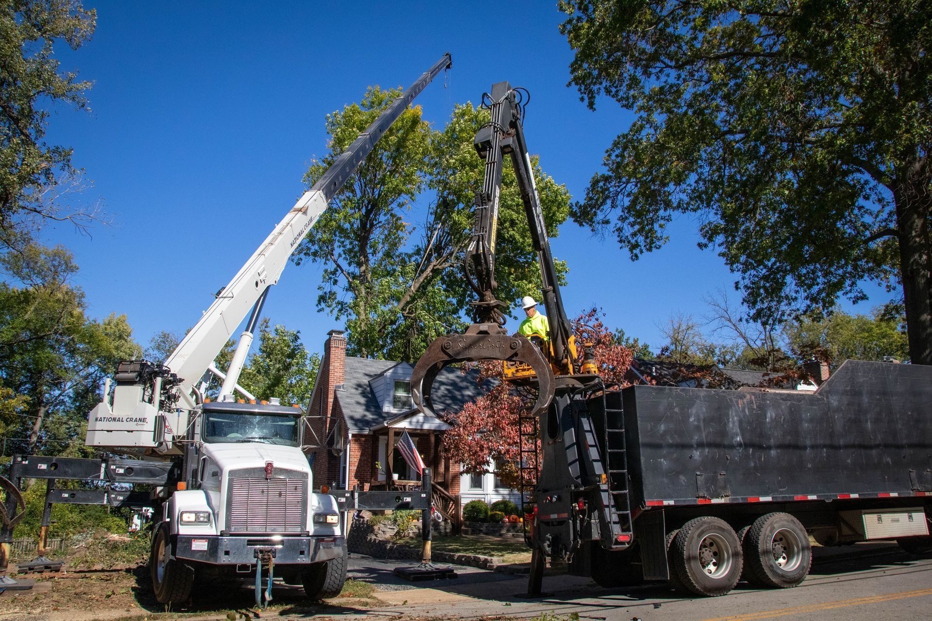 Tree removal using a crane and truck. Workers load debris into a dump truck, with a house and blue sky in the background.
