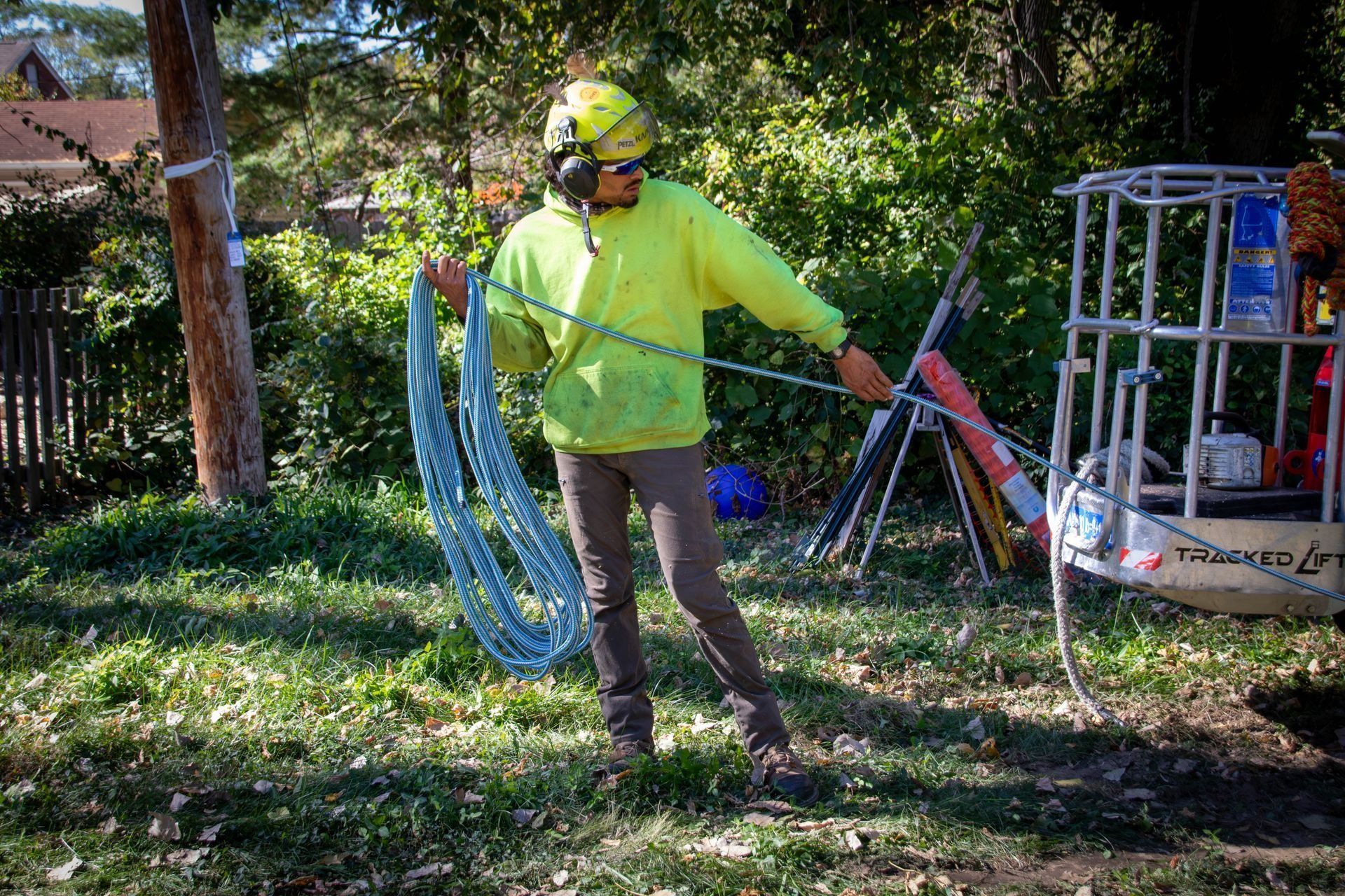 Arborist in neon yellow jacket holds blue chain near utility pole and lift, working outdoors.
