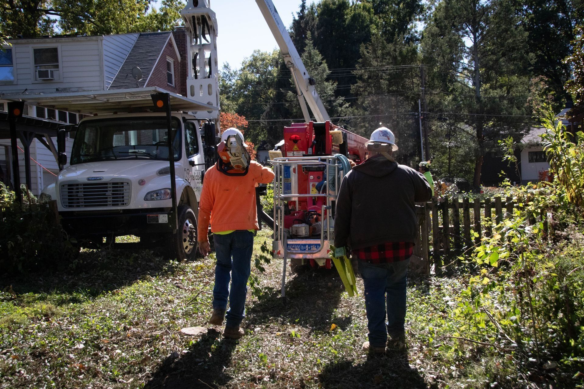Two utility workers approaching a boom lift truck near a residential area, under a sunny sky.