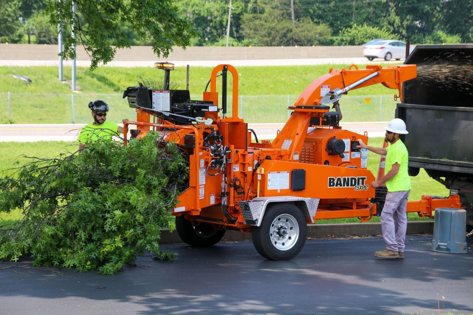 Two workers feeding tree branches into an orange Bandit wood chipper on a paved lot.