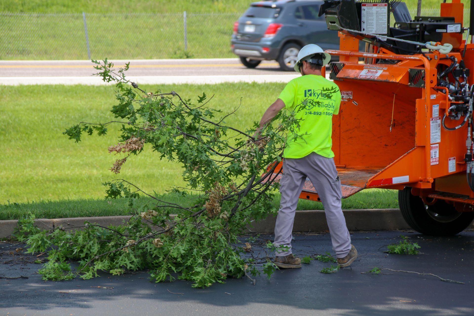 Man in safety gear feeds tree branches into a wood chipper on asphalt near a grassy area.