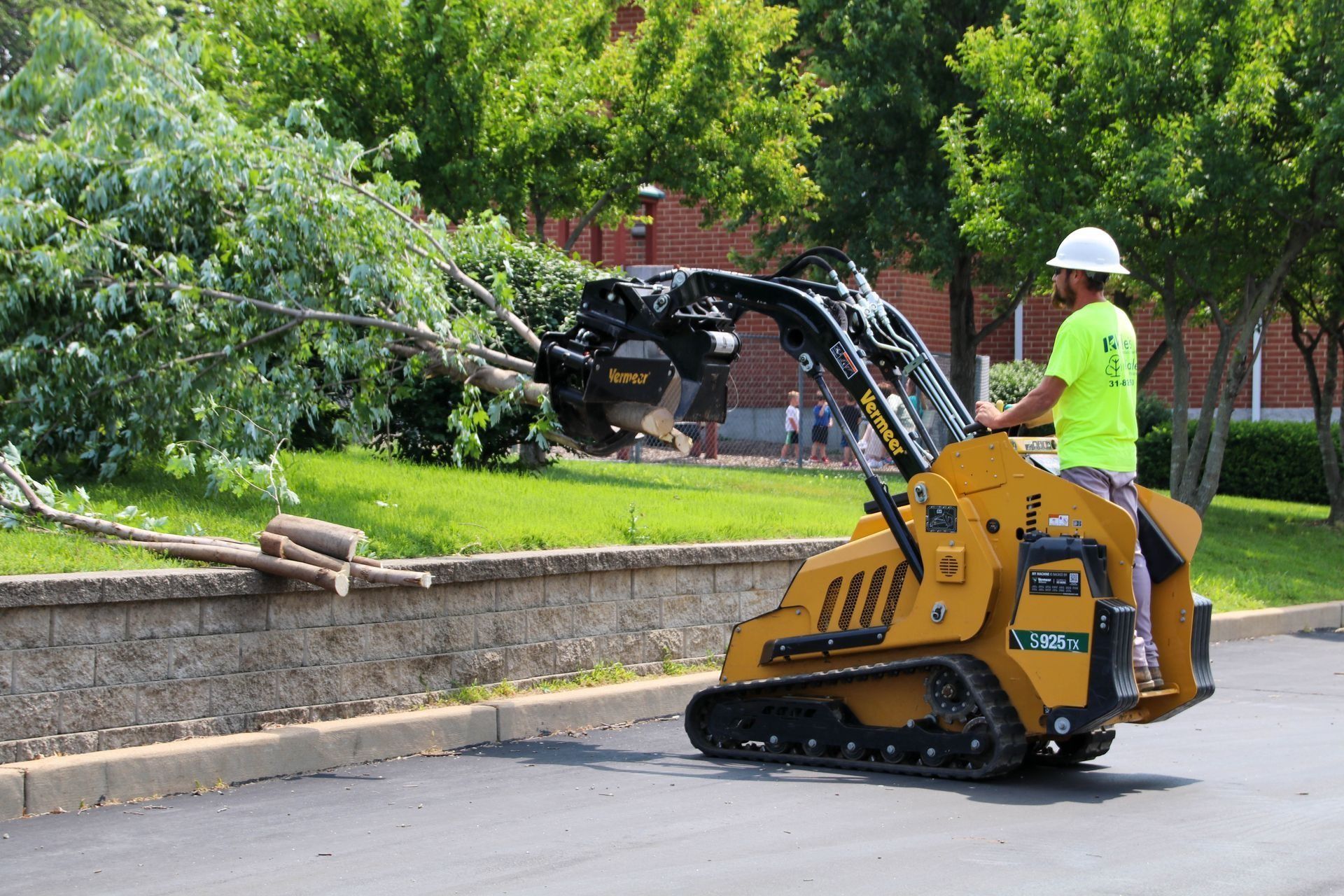 Man in neon shirt operating a yellow skid steer with tree-cutting attachment, removing a fallen tree branch.