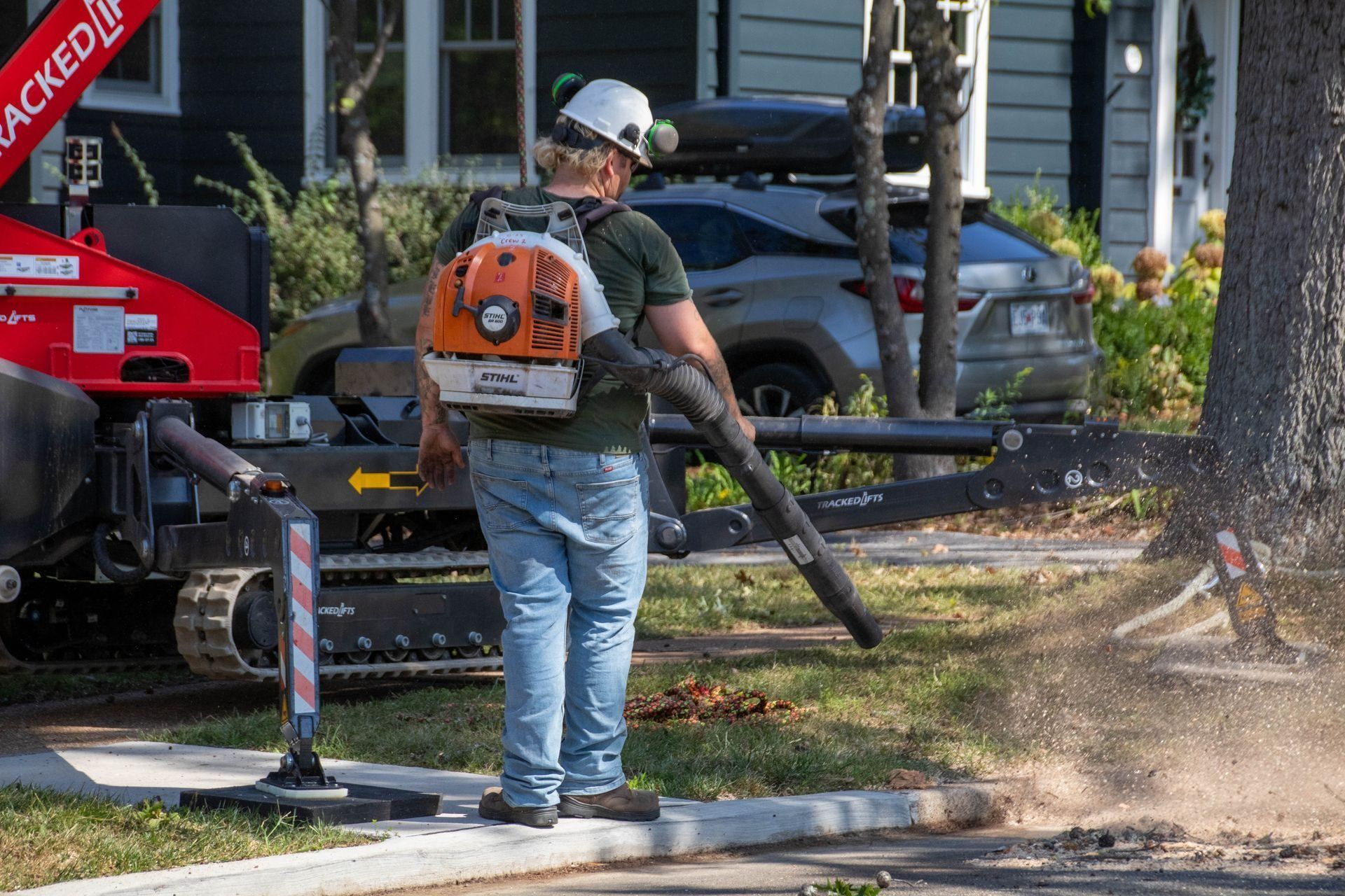 Man blowing leaves with a backpack blower near a tree and a tracked stump grinder on a residential street.