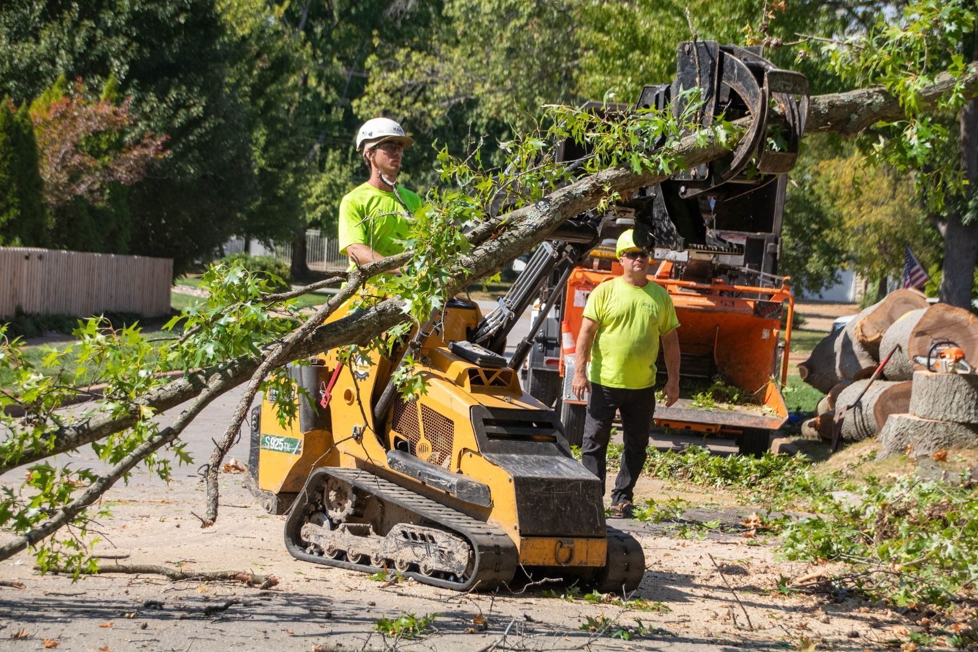 Two workers in neon green shirts using a mini track loader and wood chipper to clear tree branches on a street.