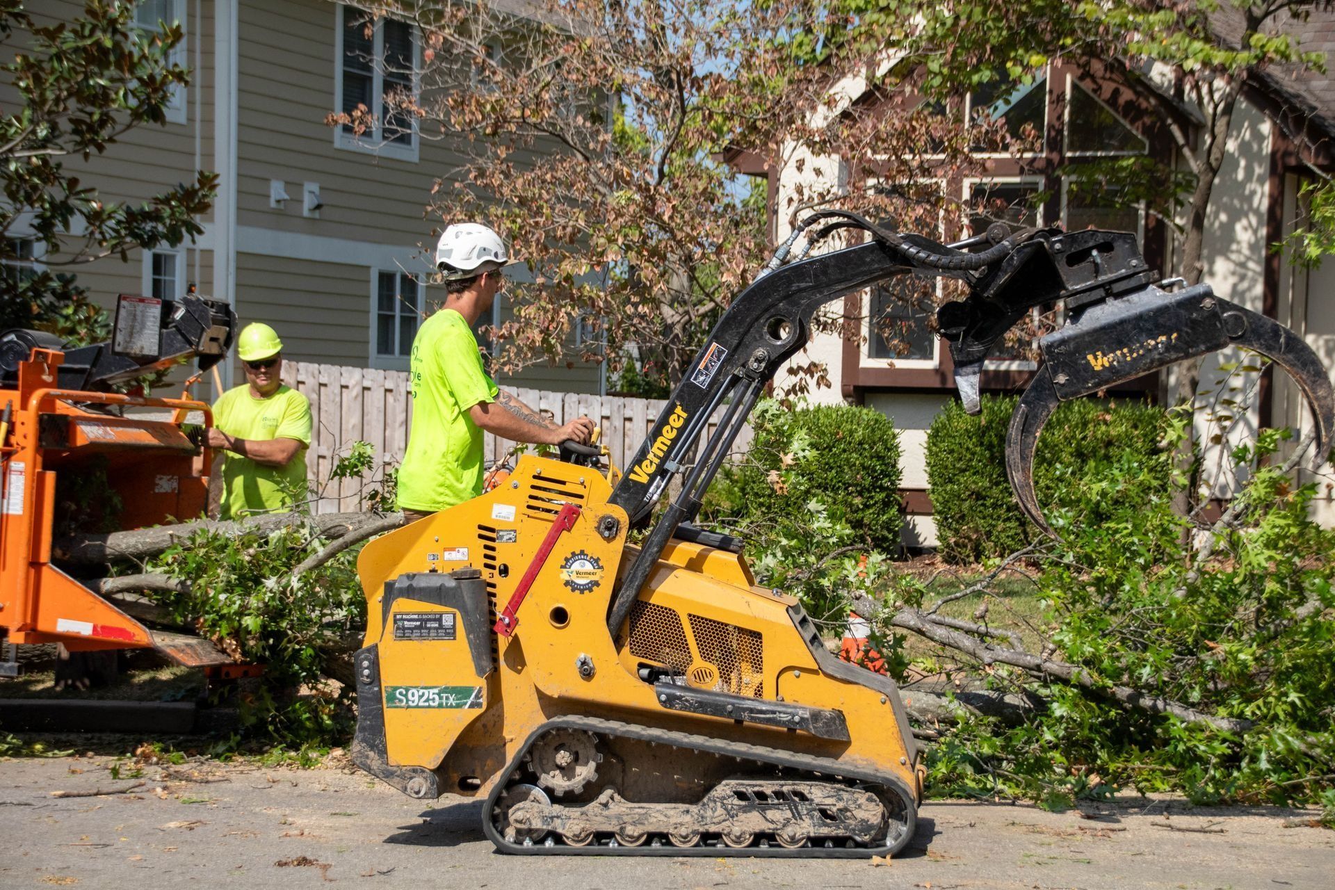 Man operating a small yellow tracked machine with a tree branch grabber, two workers, and a wood chipper in a residential area.