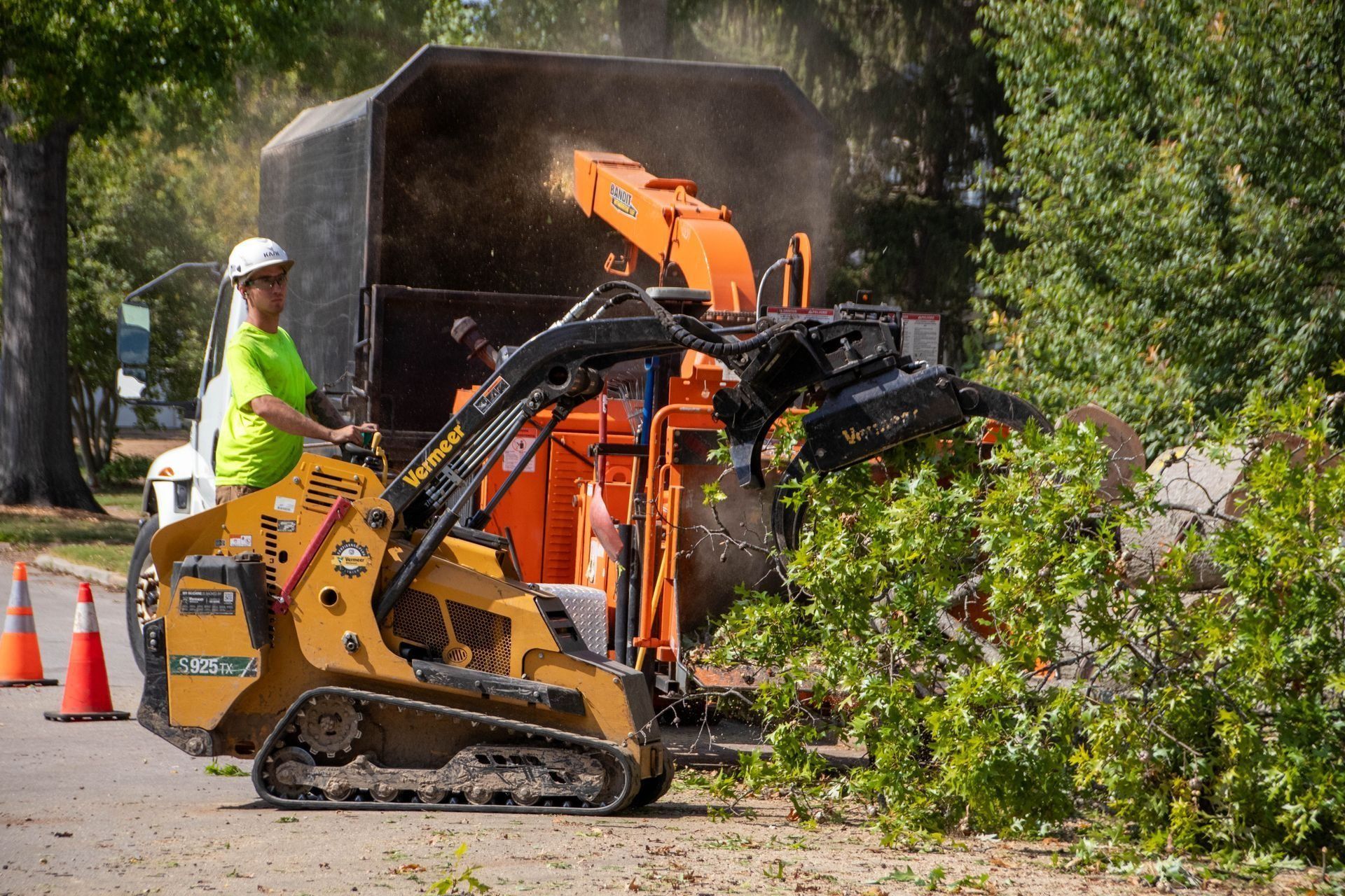 A worker in a helmet operates a small skid steer feeding a wood chipper on a street with orange cones.